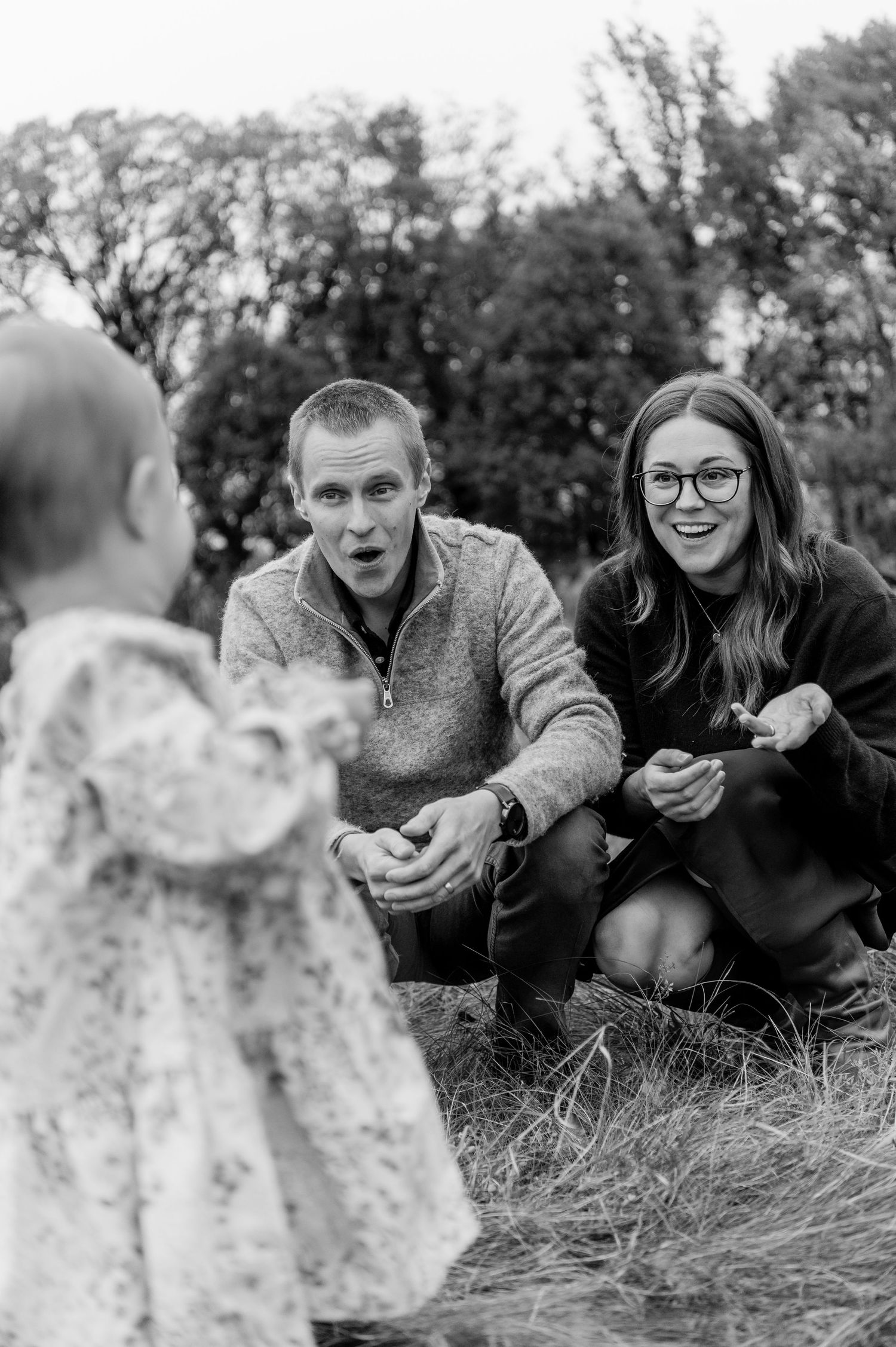 A couple crouches and reacts with delight as their toddler walks toward them outdoors. Lake Elmo, Minnesota.
