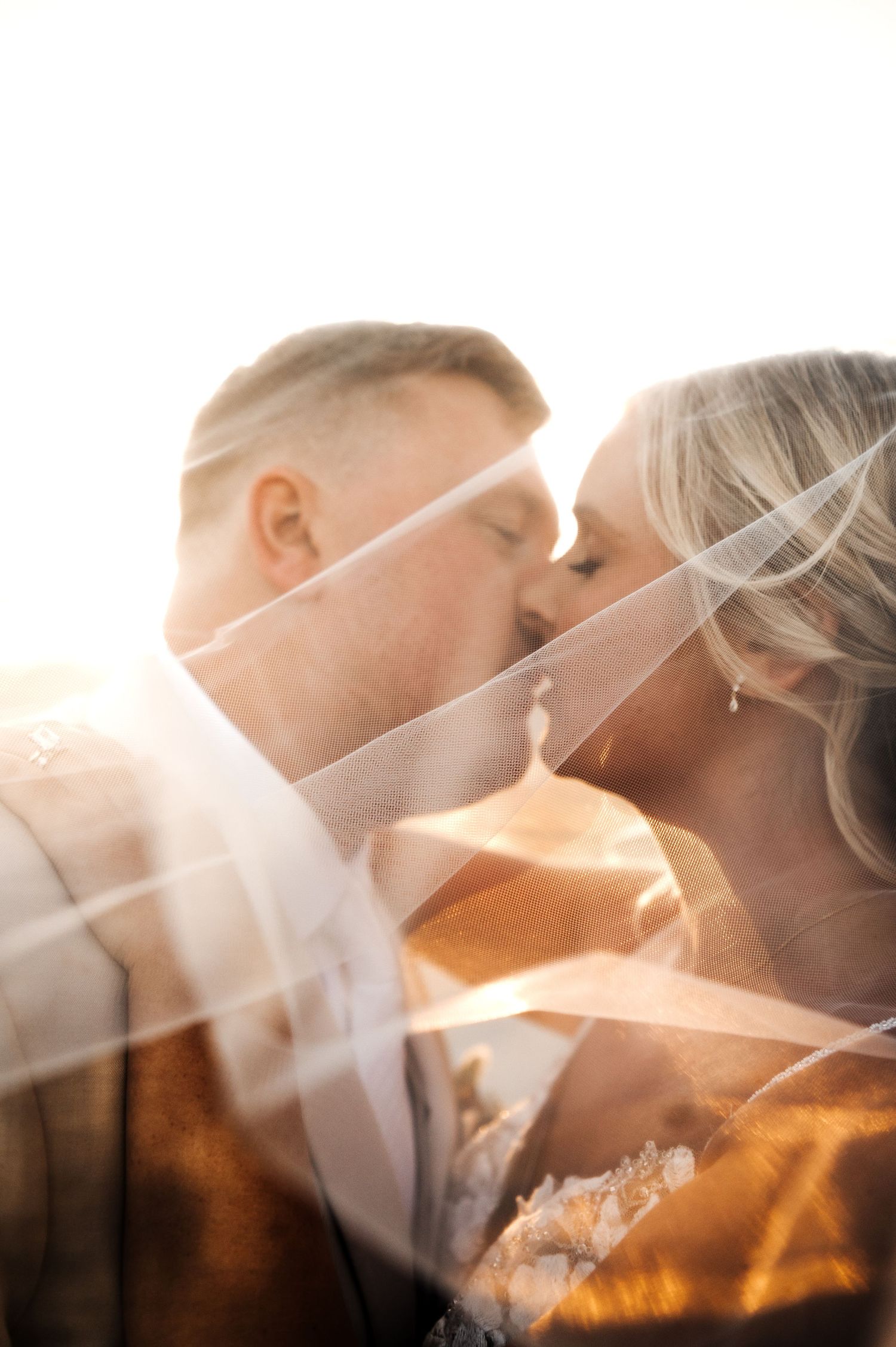 Bride and groom share romantic kiss beneath flowing veil in golden backlit wedding portrait. Wisconsin. Minnesota.