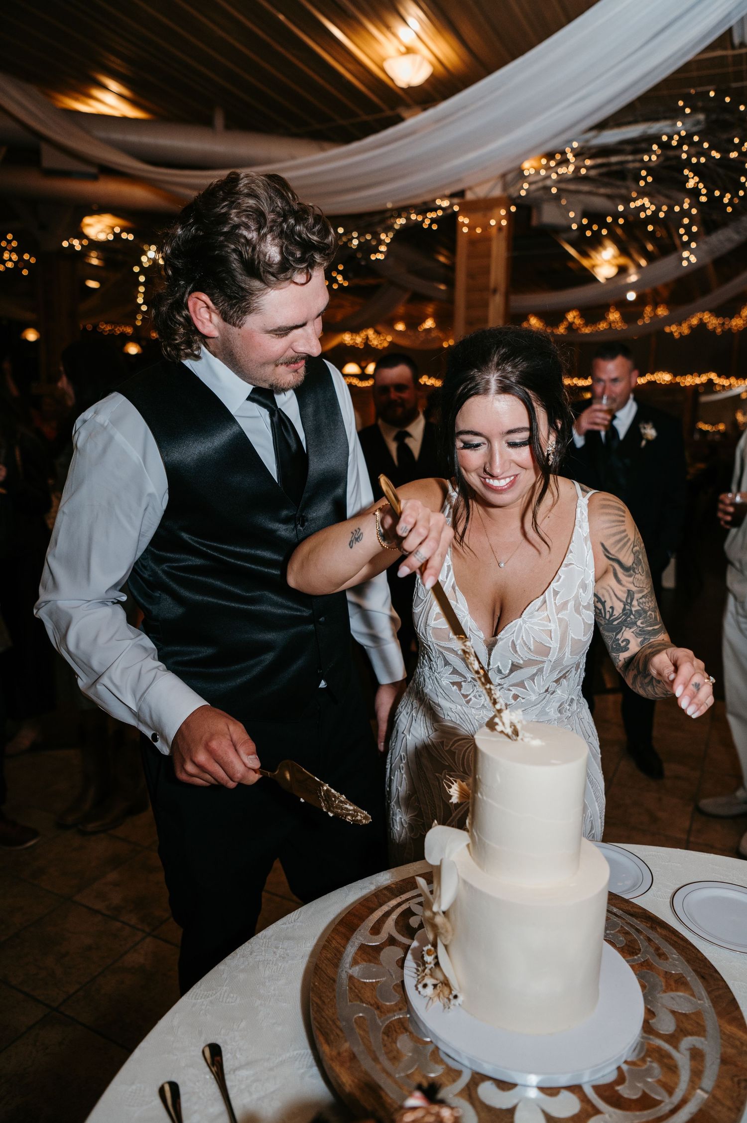 Bride and groom cutting white tiered wedding cake at reception with string lights.