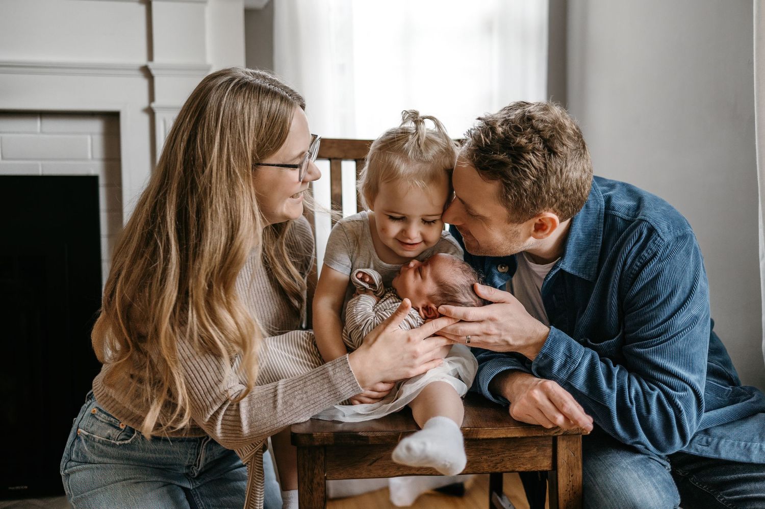 Two parents smile as their toddler holds a newborn baby on a wooden chair.