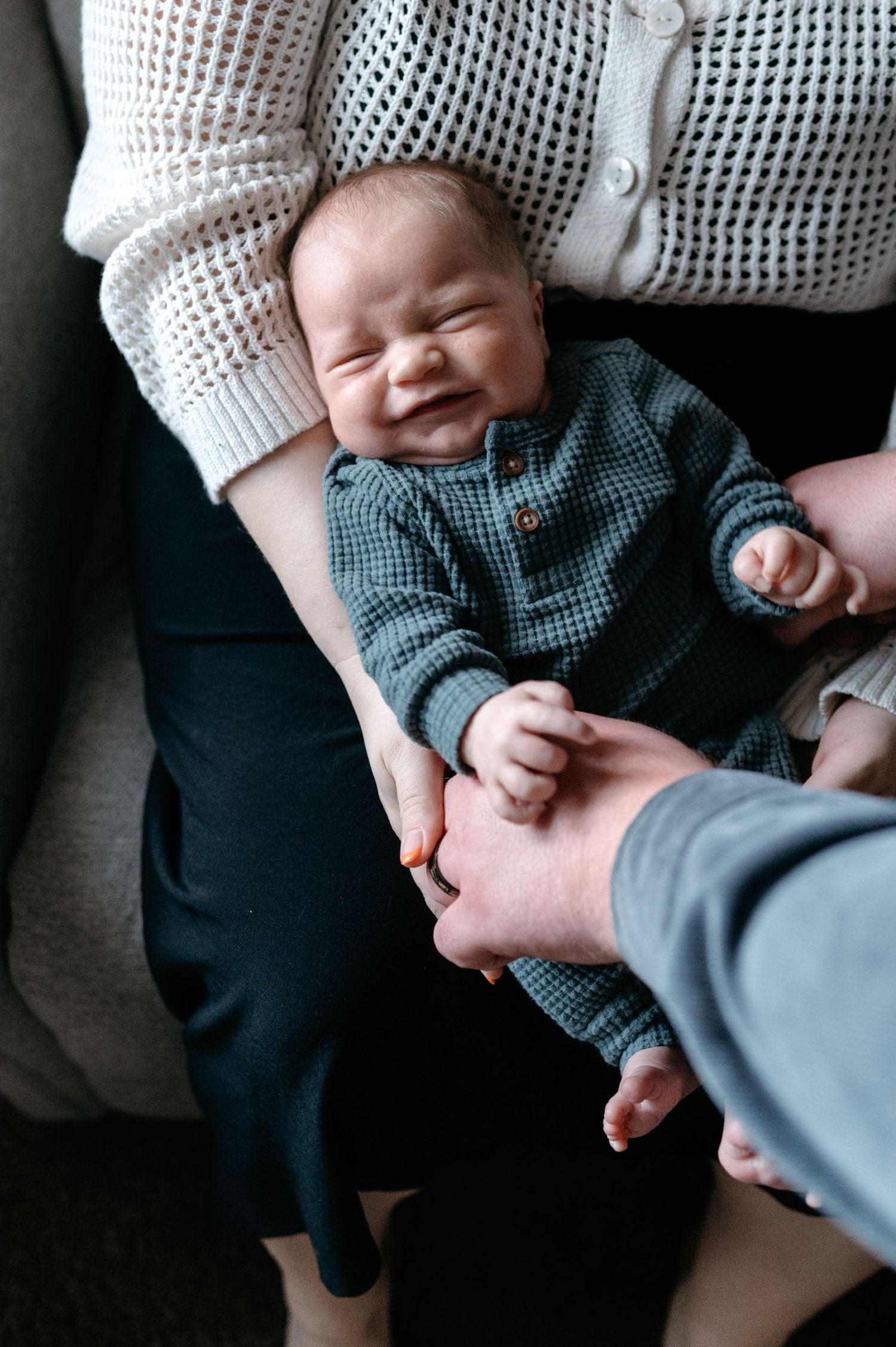 Smiling newborn baby in green checkered outfit being held by parents on sofa.
