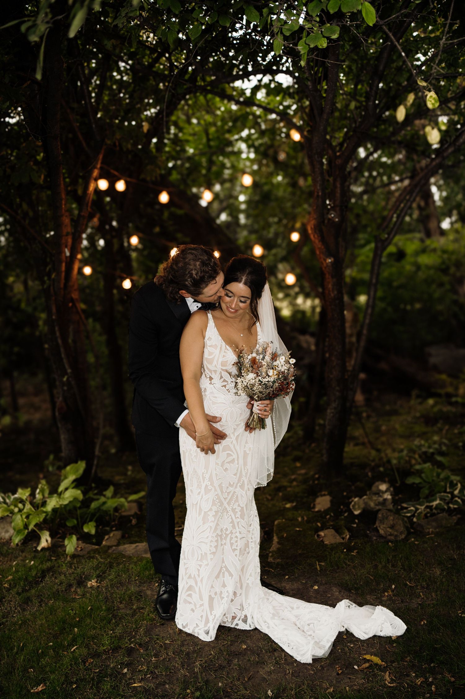 Bride and groom embracing under Edison bulb lights in lush garden at dusk. Wisconsin. Minnesota.