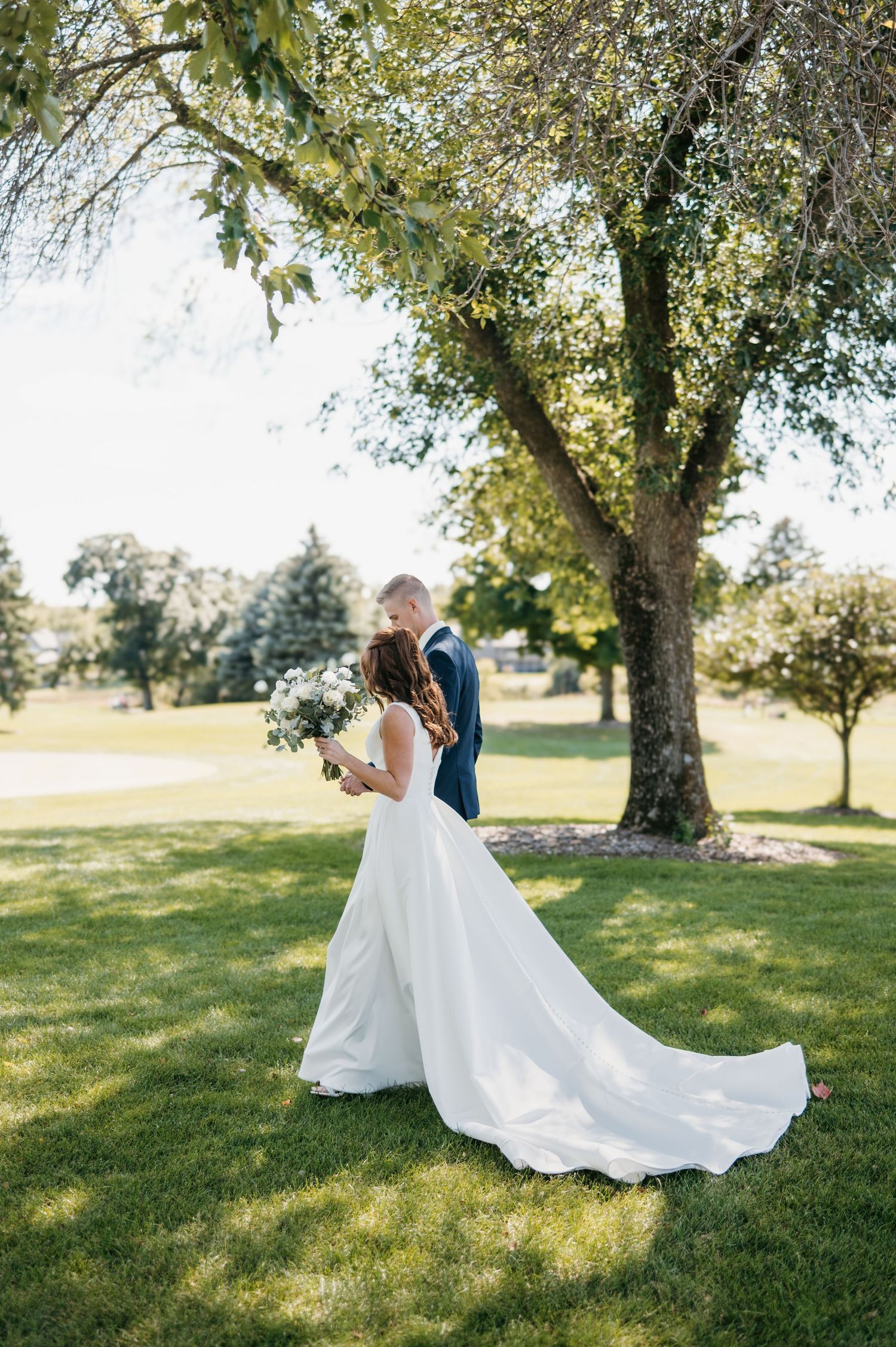 Bride in flowing white gown and groom embracing under large tree on sunny day.