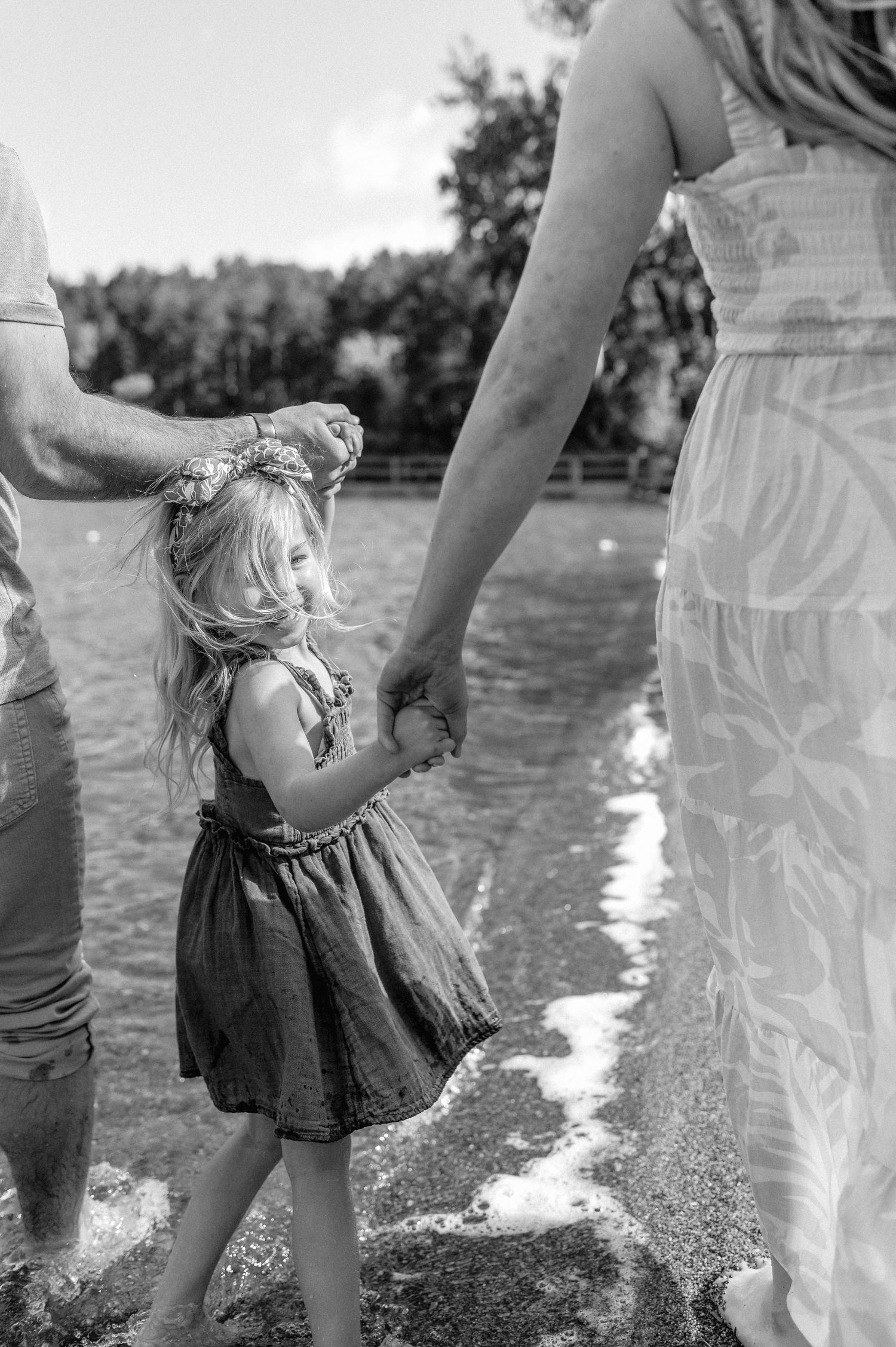 Black and white photo of young girl holding parents hands while wading in lake.