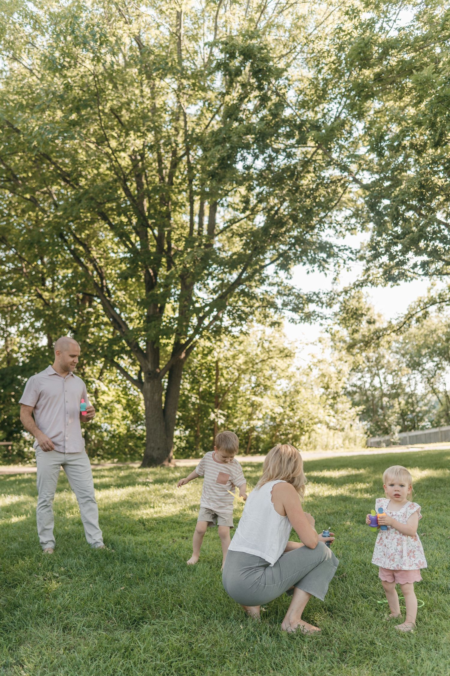 Family of four playing with bubbles on green grass in sunlit park with tall trees.