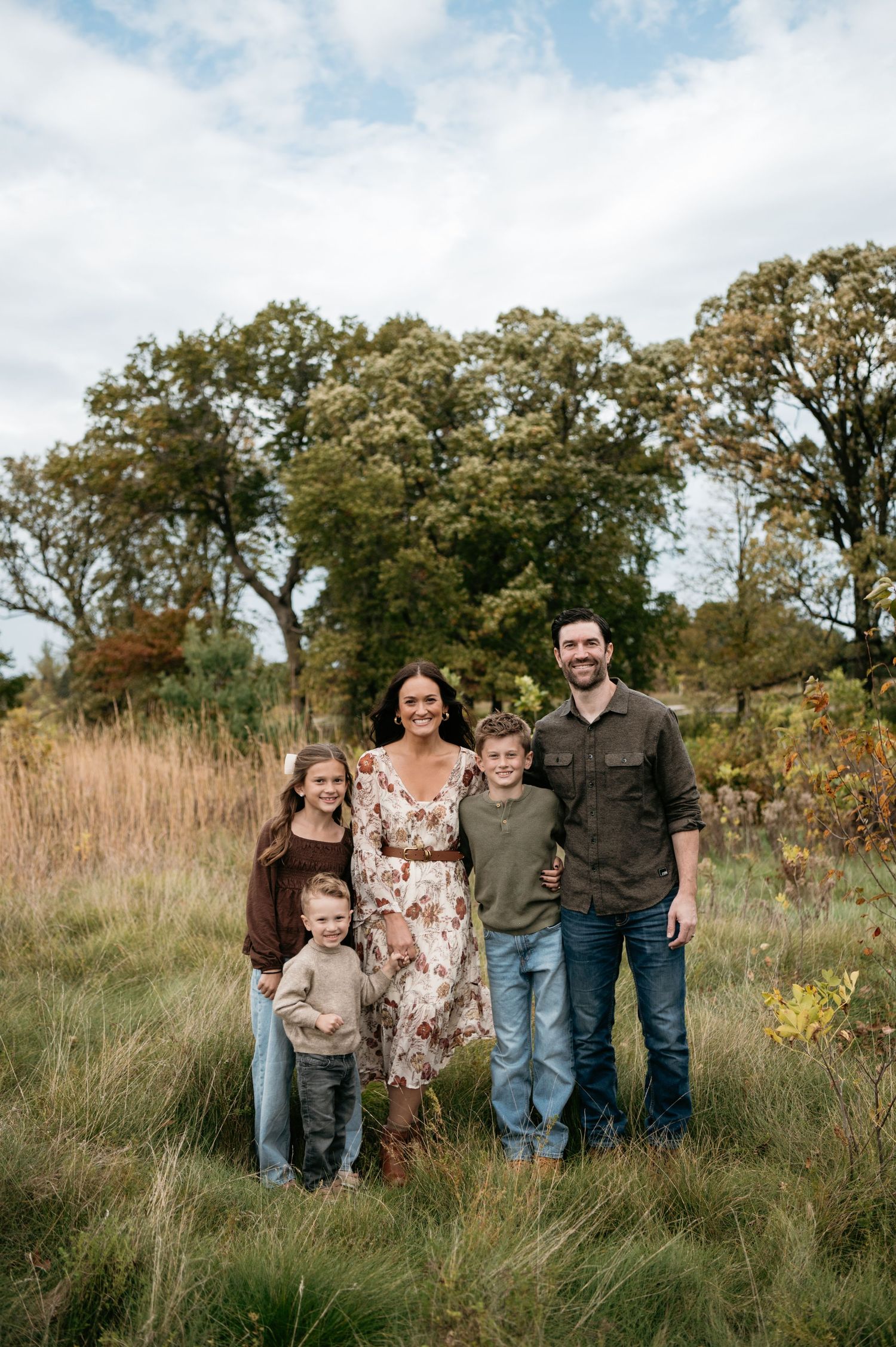 A family of five poses together in a scenic fall field with colorful autumn trees in the background. Woodbury, Minnesota.