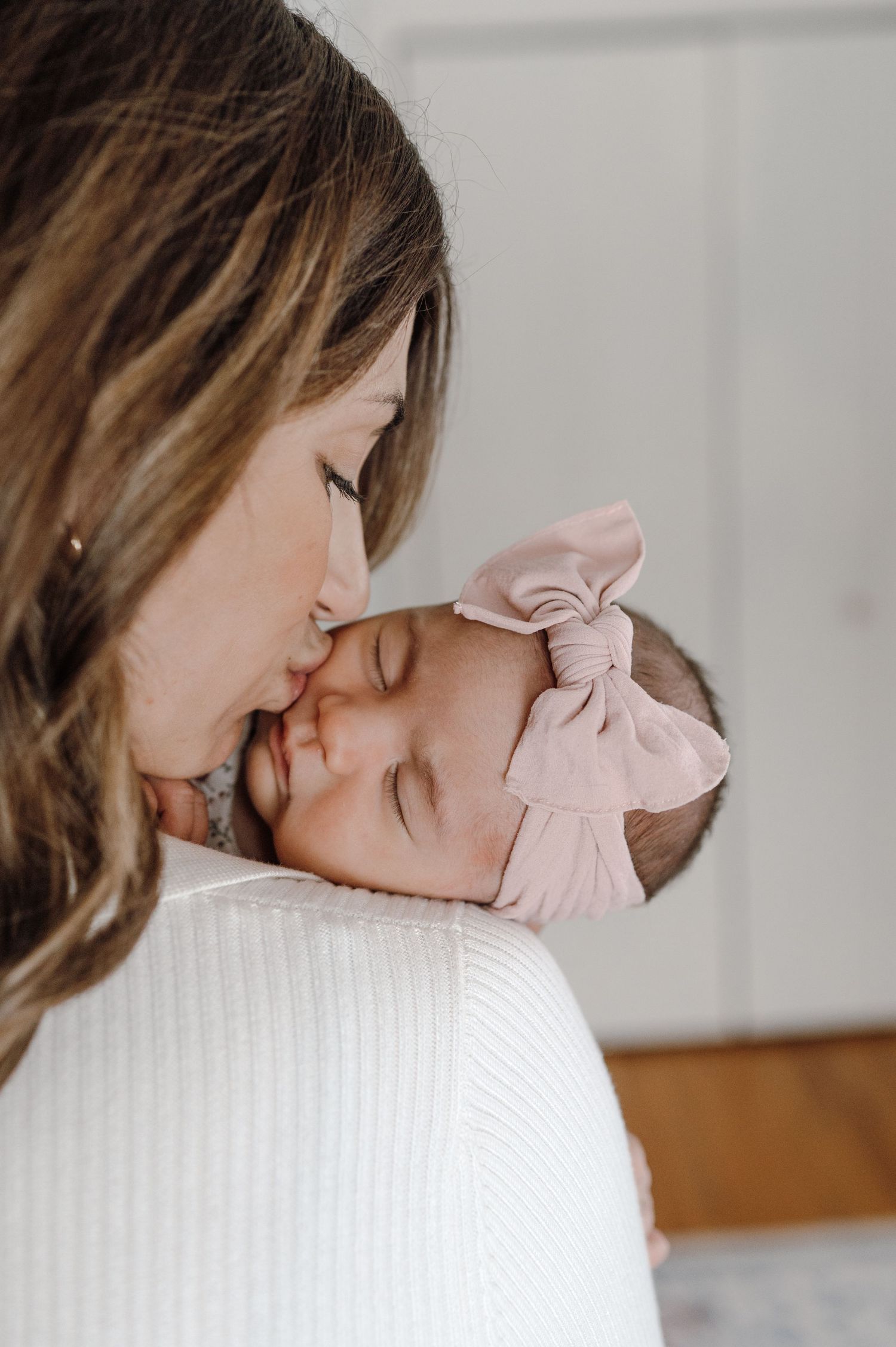 Mother gently kissing her sleeping newborn baby girl wearing a pink bow headband.