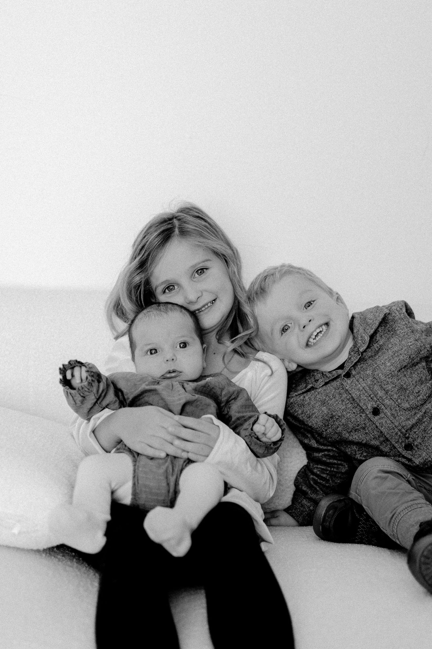 Black and white photo of two siblings smiling while holding a newborn baby on a white couch.
