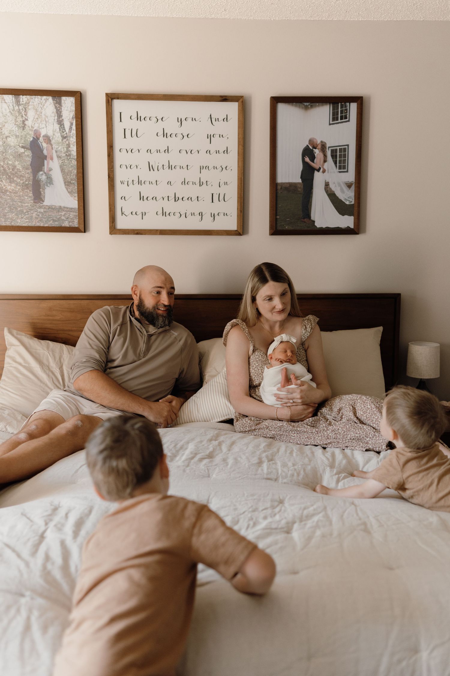 Family with newborn baby relaxing on bed beneath framed wedding photos on wall.