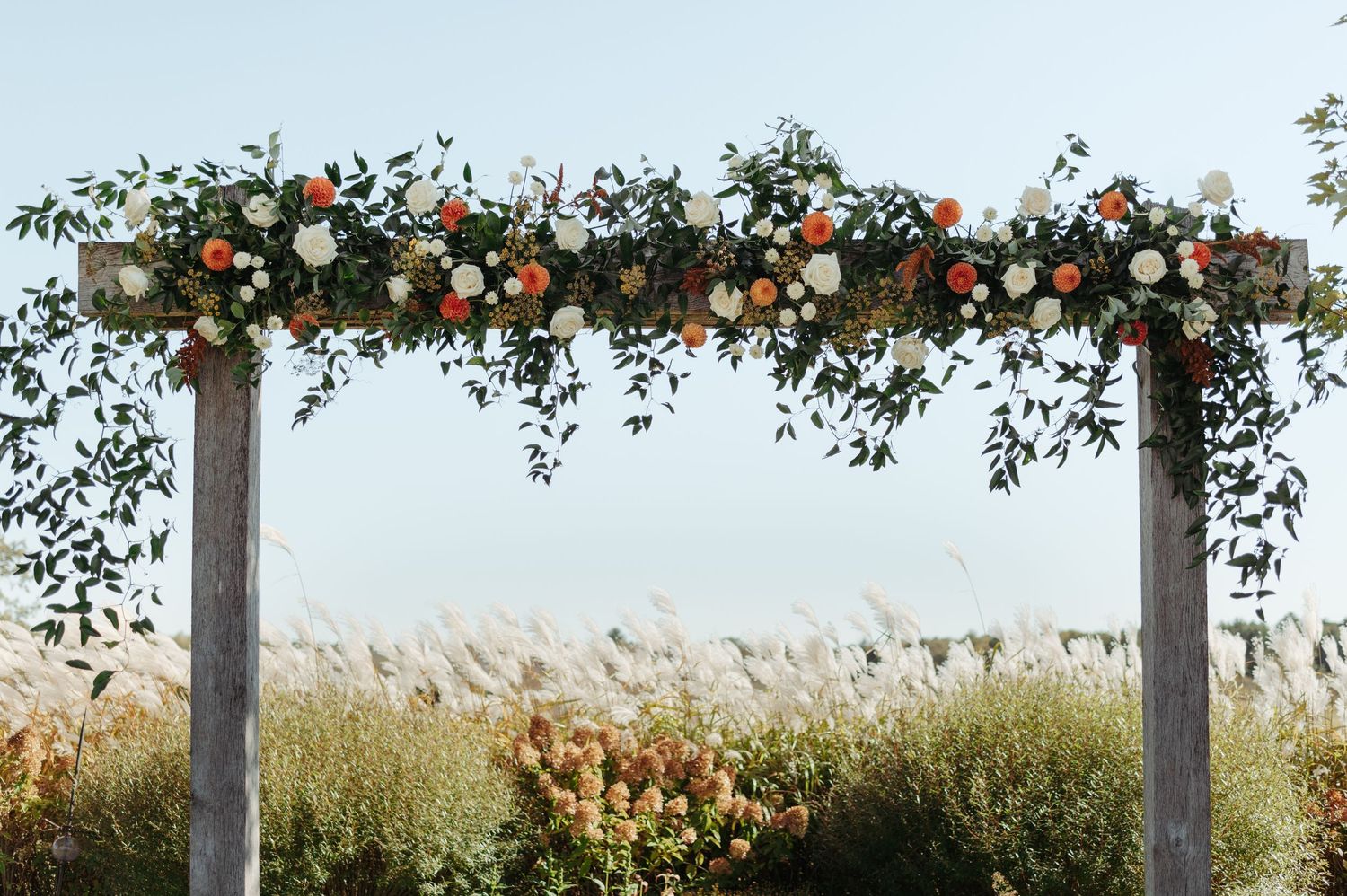 Rustic wooden wedding arch adorned with lush orange and white floral garland in open field.