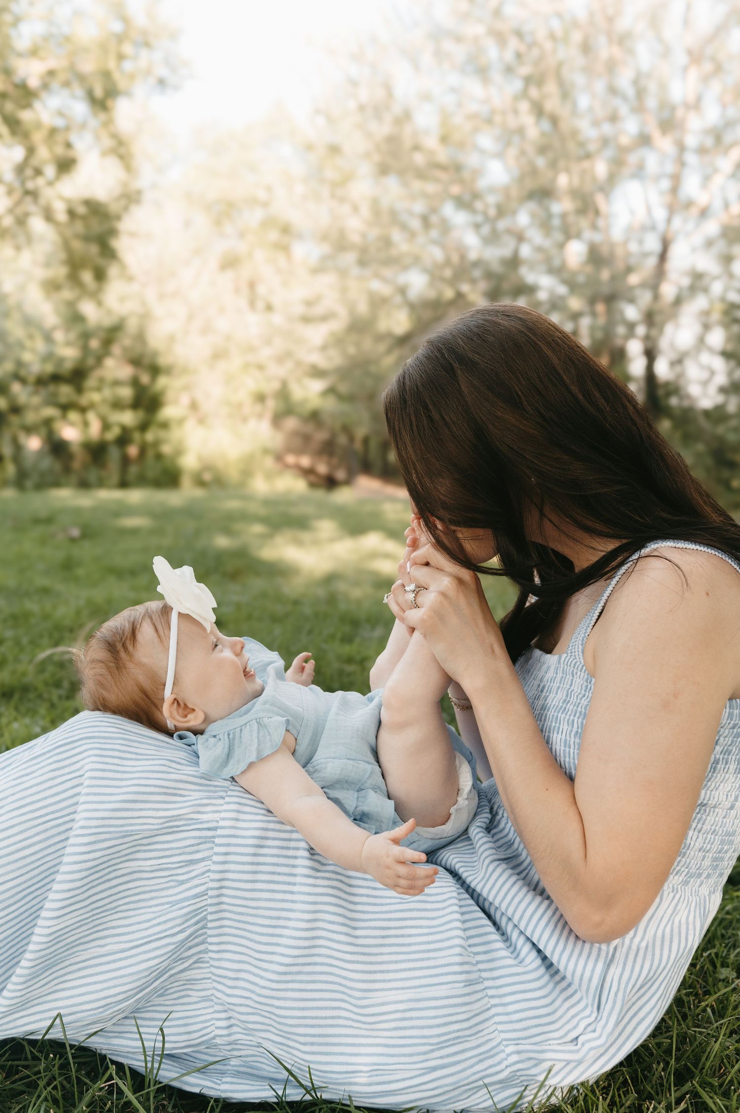 Dark-haired mother in striped dress playing with baby girl lying on blanket in sunny park.
