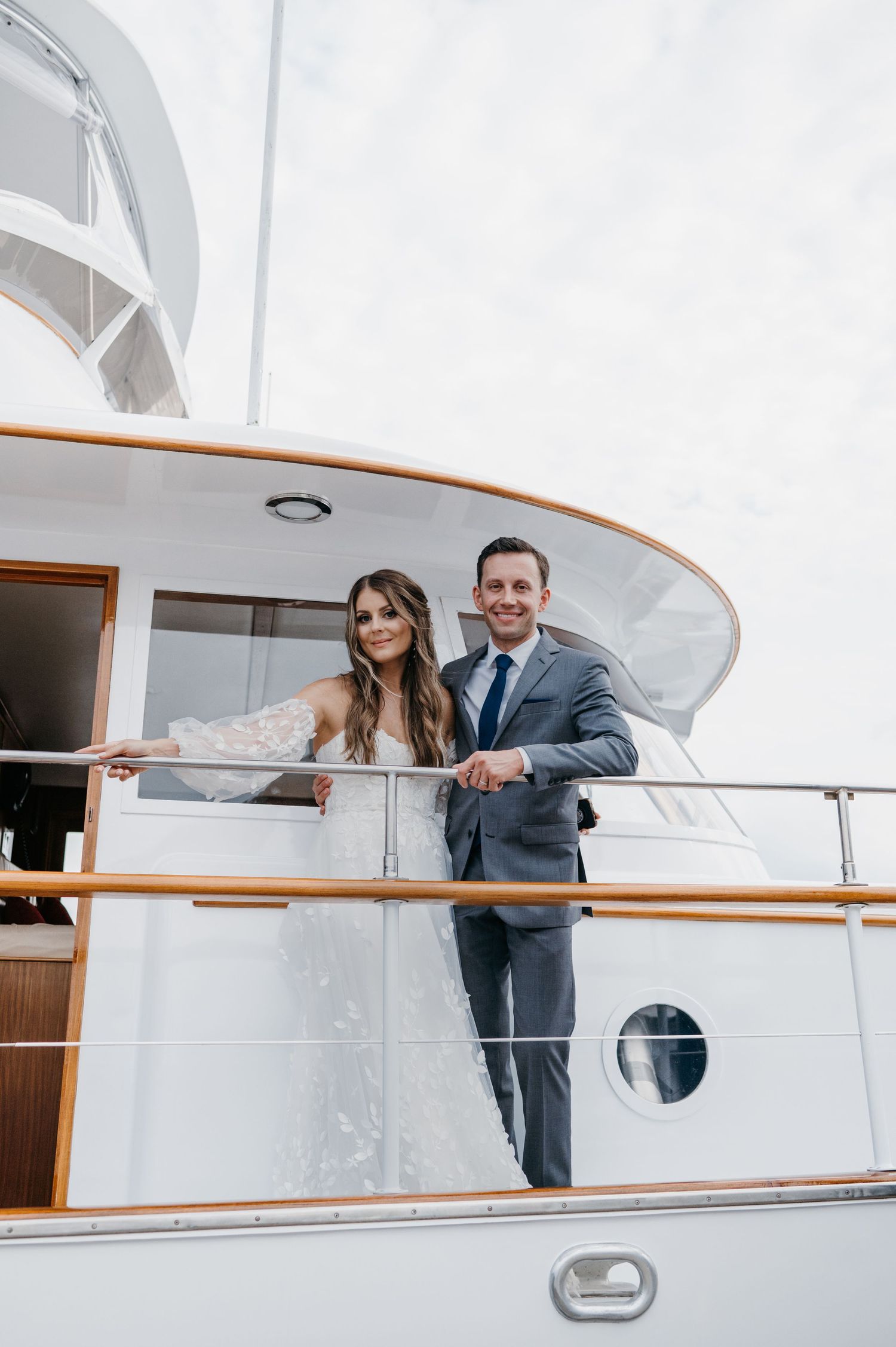 Smiling bride and groom standing on upper deck railing of white luxury yacht.