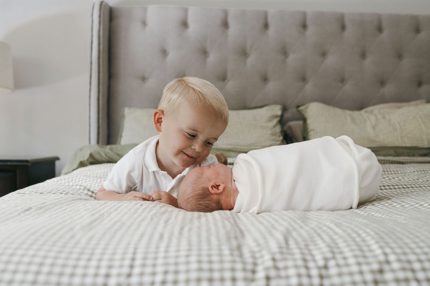 Adorable toddler boy smiling at swaddled newborn sibling on a cozy bed. Wisconsin. Minnesota.