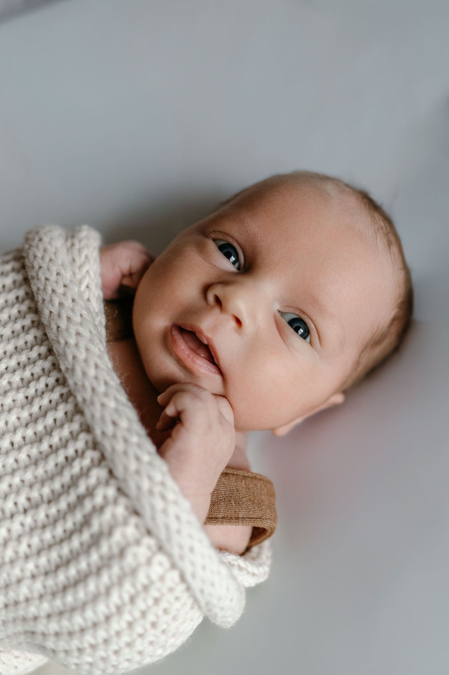 Alert newborn baby in beige knit wrap with finger in mouth gazing upward.