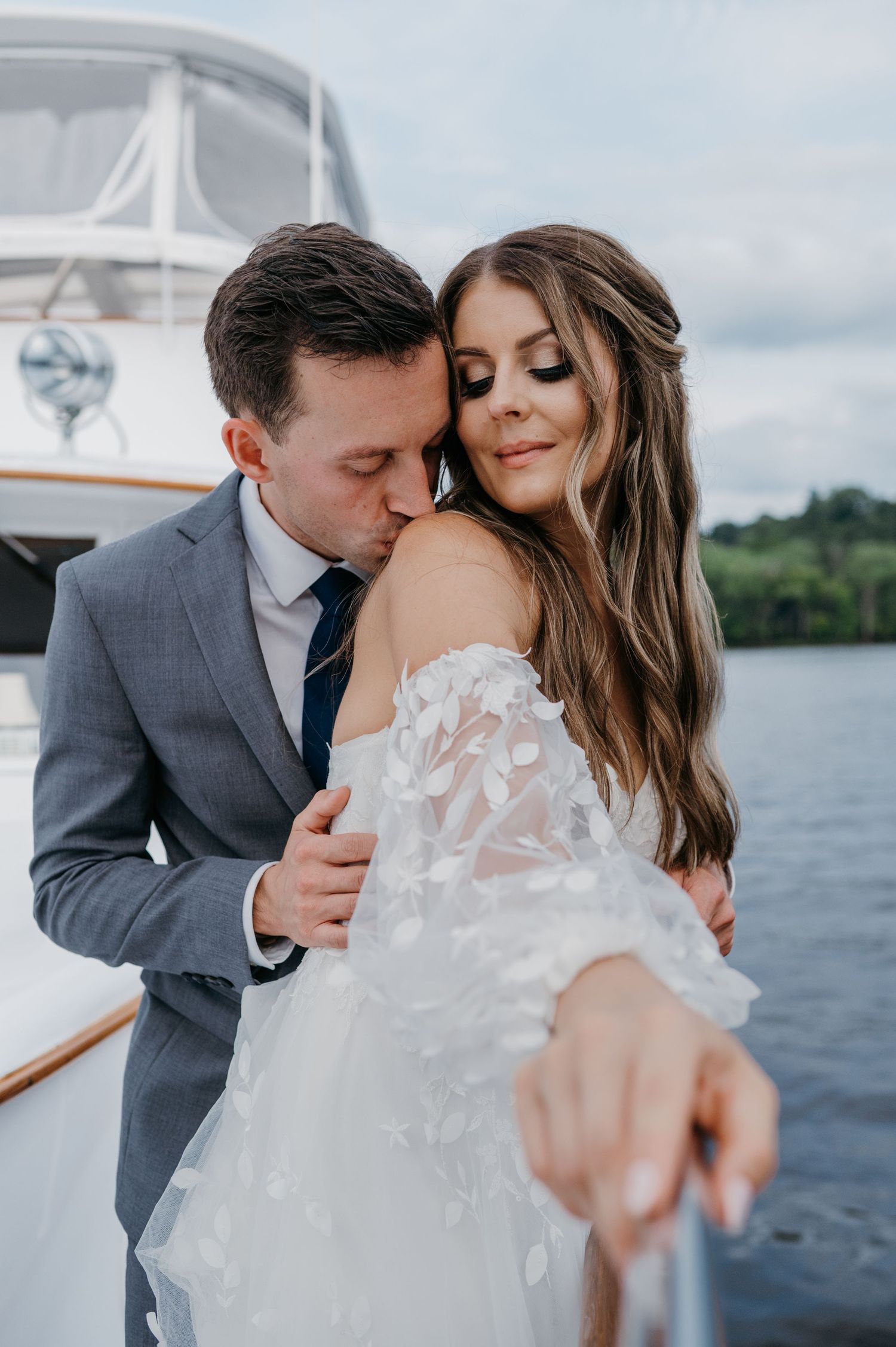 Groom in grey suit kissing bride in floral off-shoulder gown on yacht deck.