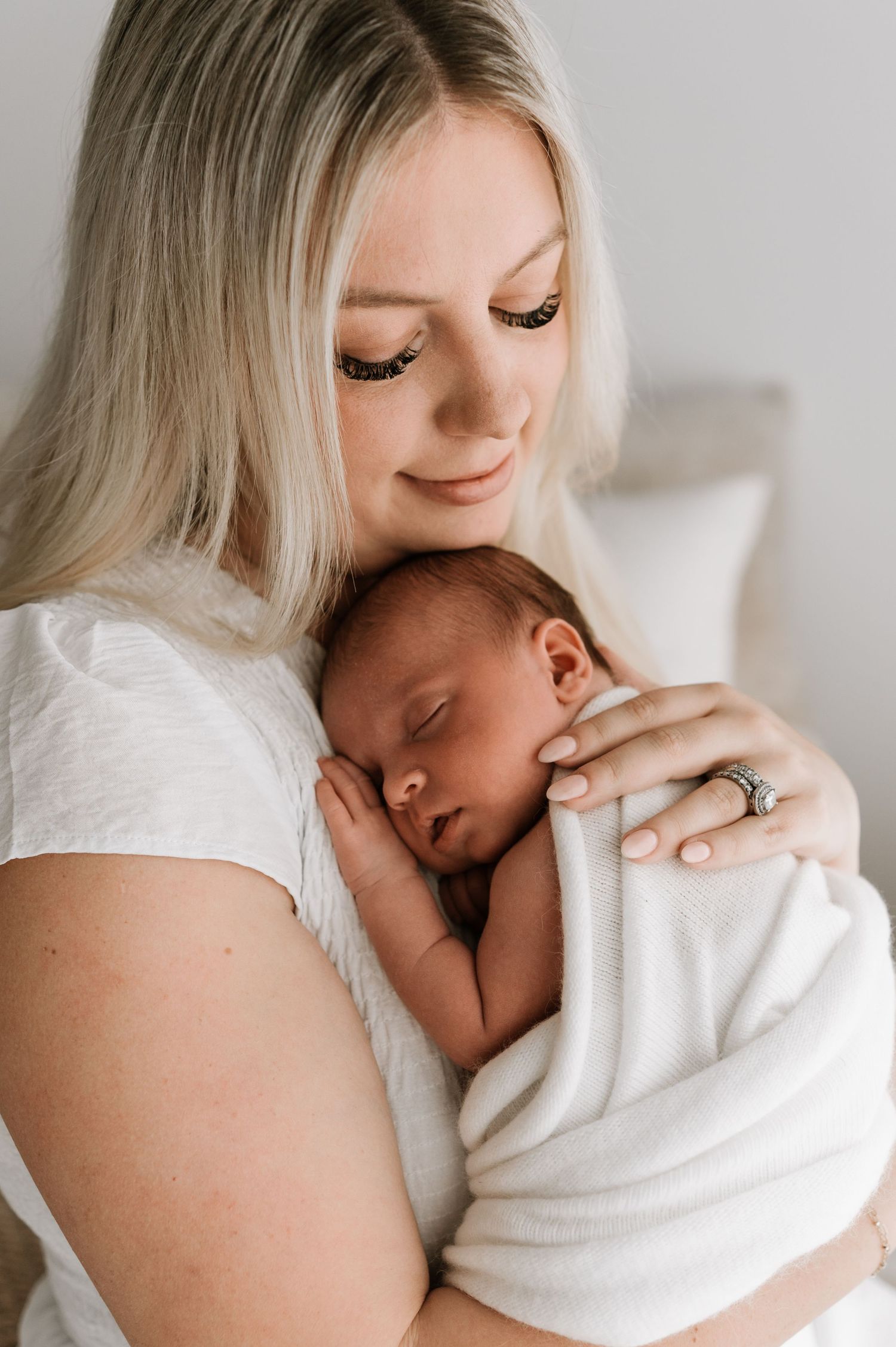 Blonde mother tenderly holding sleeping swaddled newborn baby against her chest.