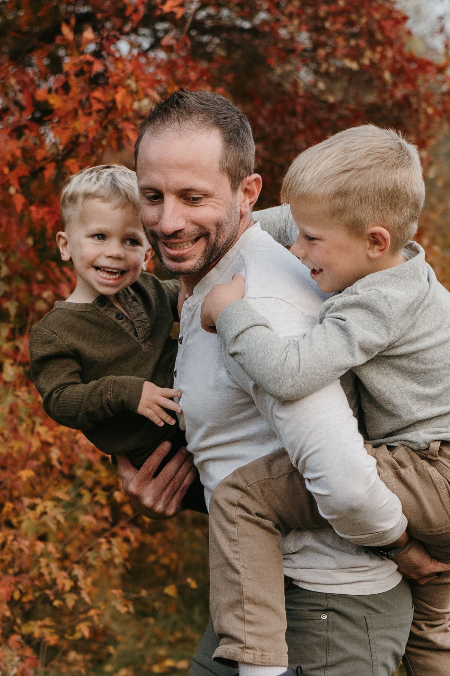 A smiling father holds two young blonde boys in front of vibrant red autumn foliage. Woodbury, Minnesota.
