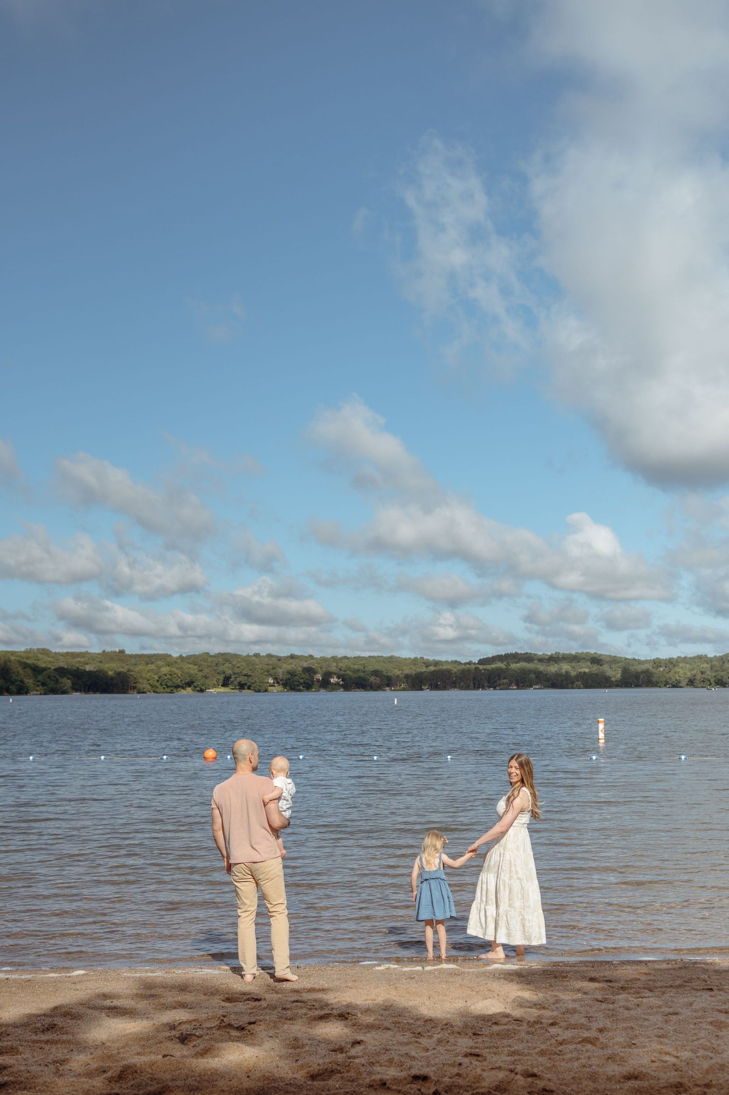 Young family of four standing at lakeside beach on sunny day with blue sky and clouds.