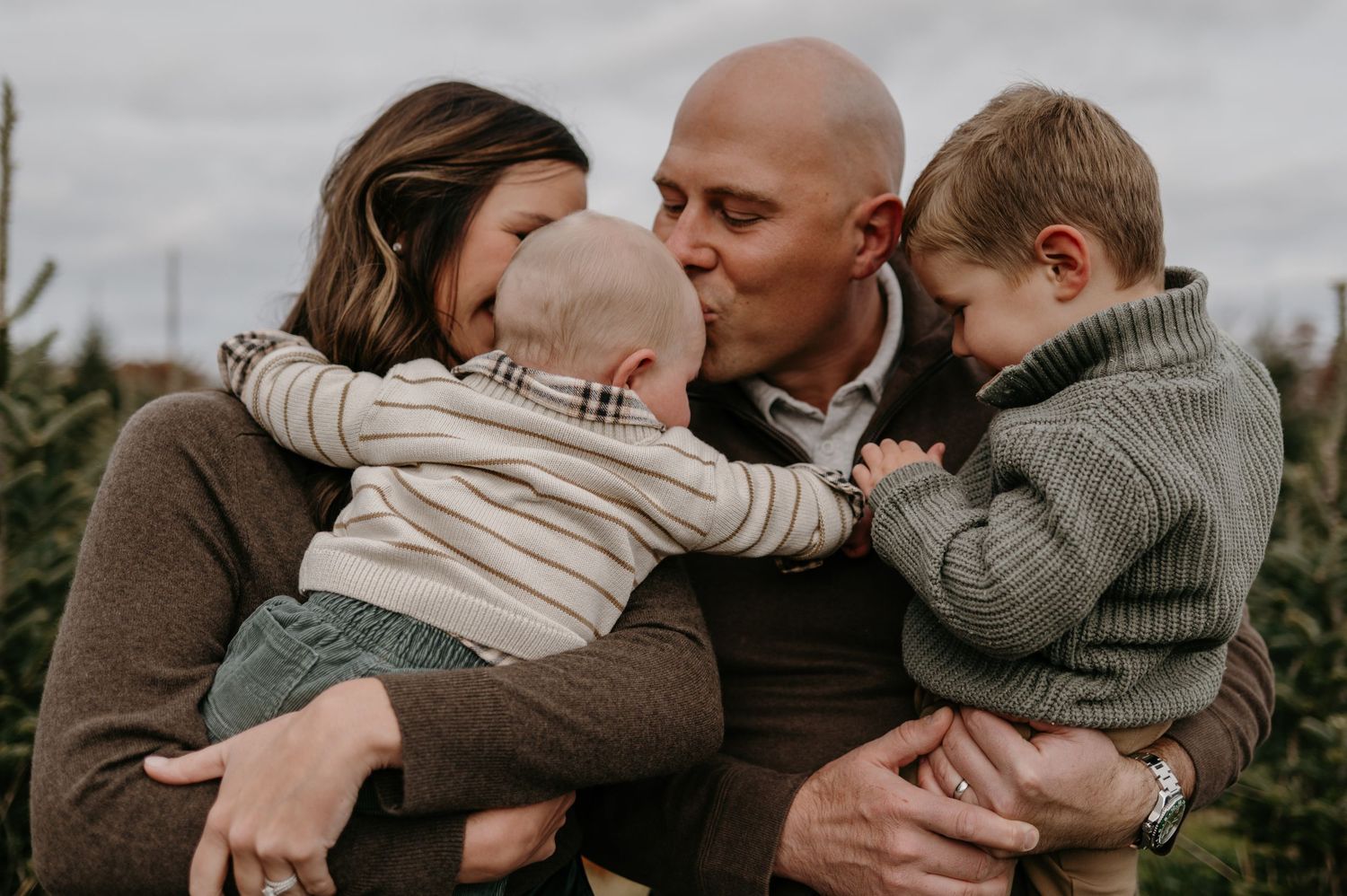 Loving family moment with parents kissing baby while toddler cuddles close outdoors. Lake Elmo, Minnesota.