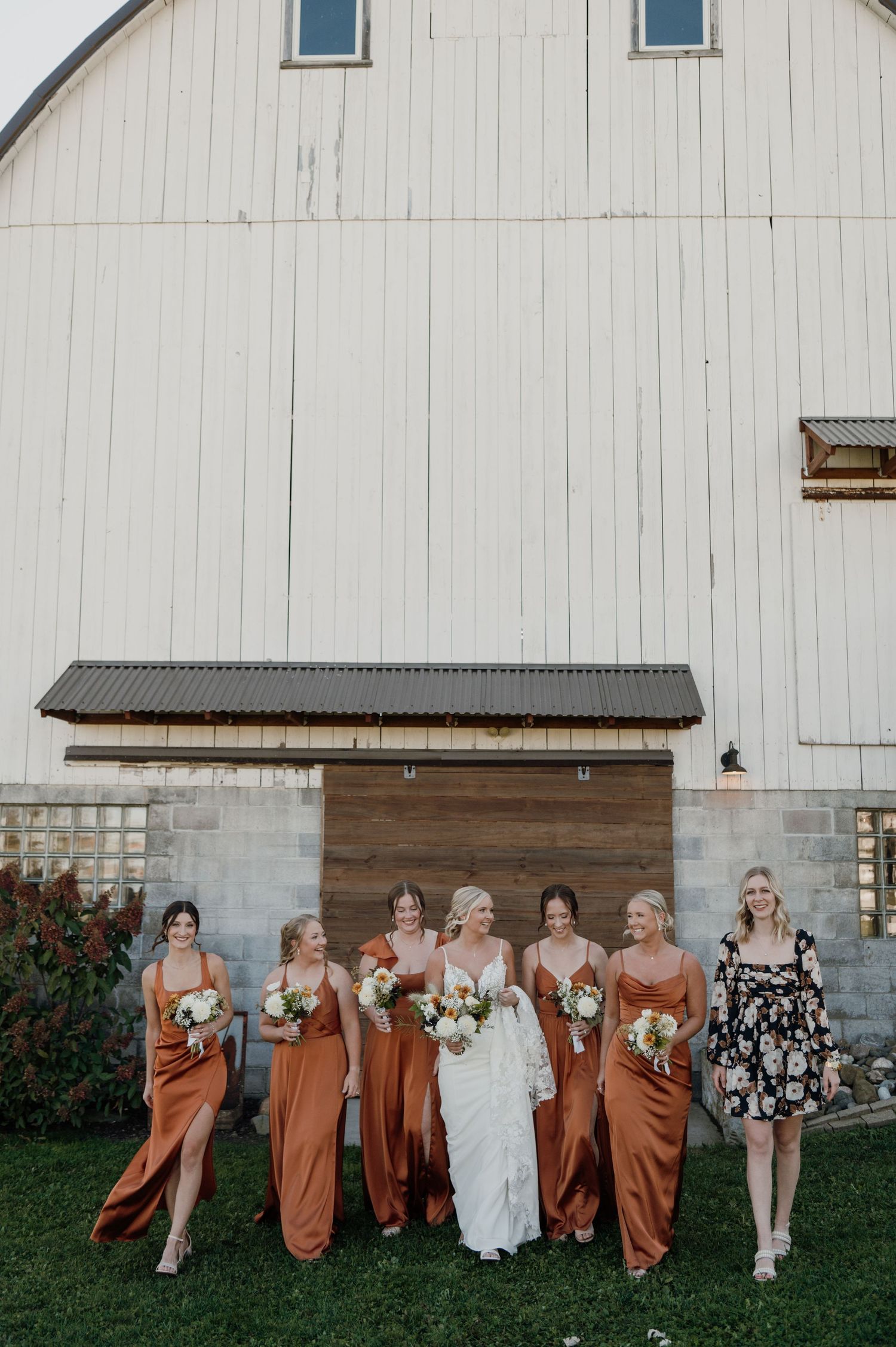 Bride and bridesmaids in rust orange satin dresses holding white bouquets posing in front of a white barn wedding venue.