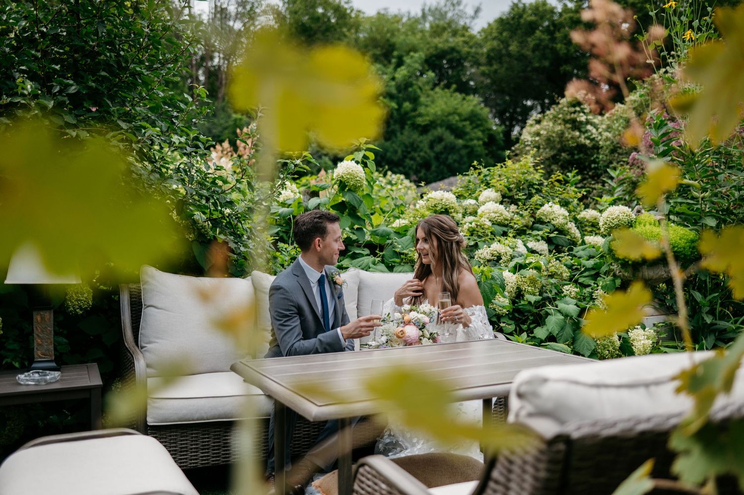 Bride and groom toasting champagne at outdoor garden table surrounded by lush greenery.