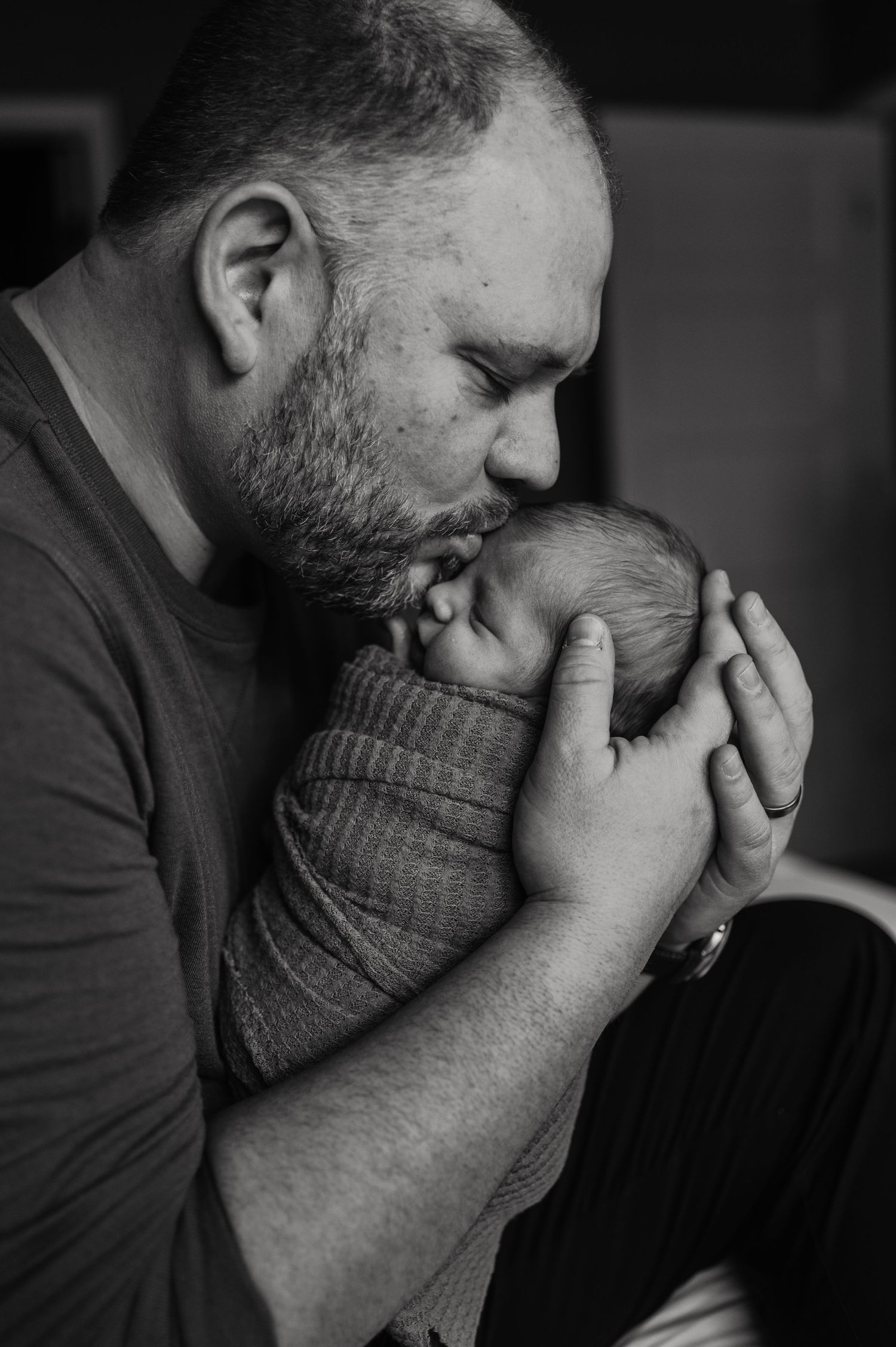 Black and white portrait of a father tenderly kissing his swaddled newborn baby.