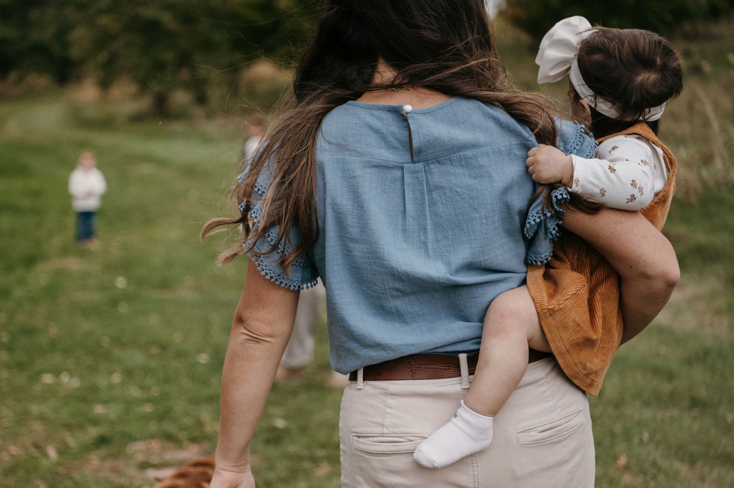 Mother carrying baby on her hip while toddler walks ahead in a grassy field. Woodbury, Minnesota.