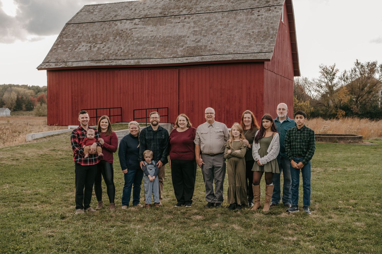 A large extended family of eleven poses together in front of a classic red barn in fall. Woodbury, Minnesota.