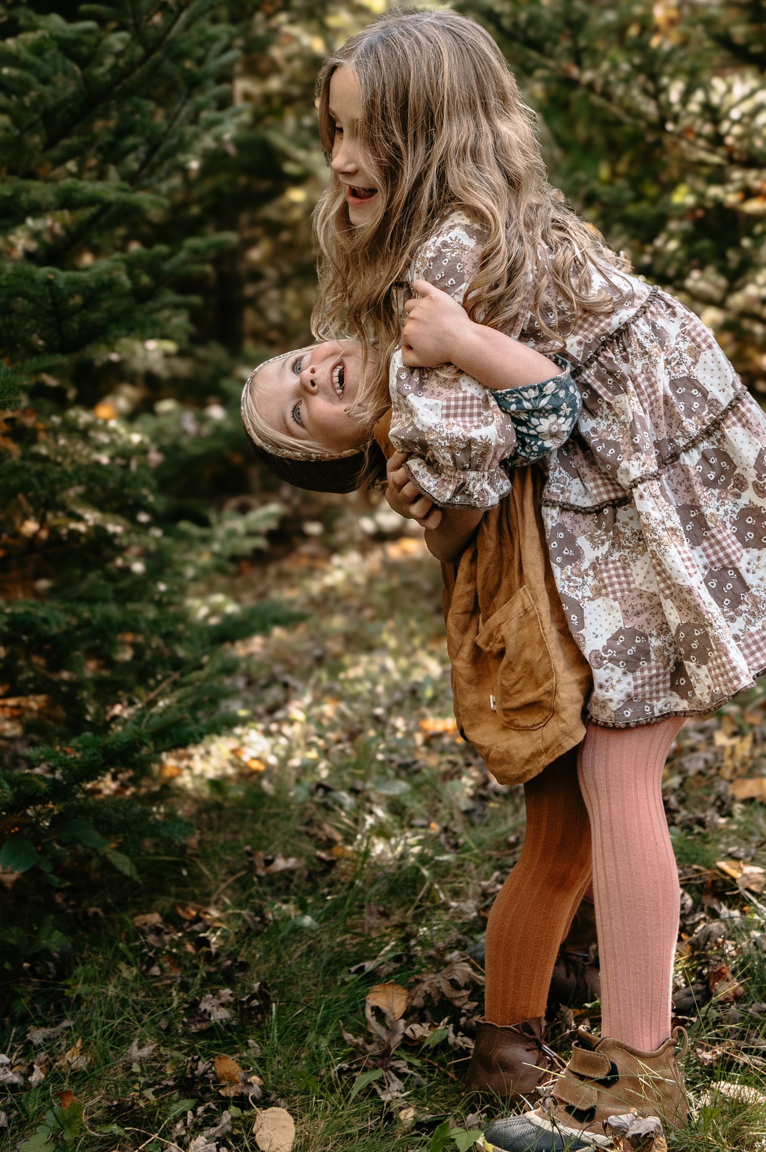 A laughing girl playfully holds a younger child upside down among evergreen trees in autumn. Woodbury, Minnesota.