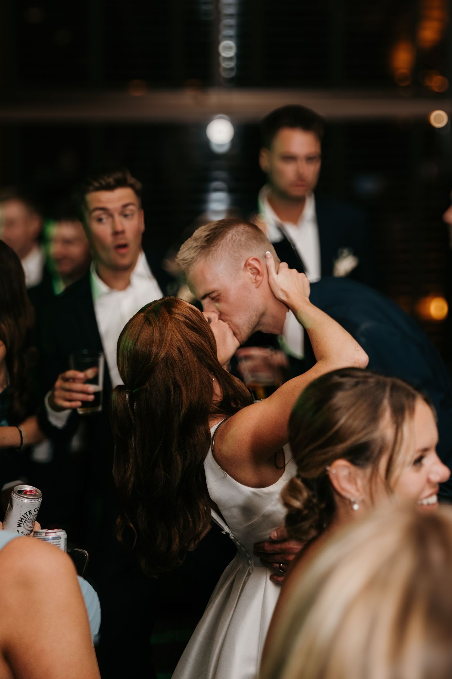 Bride and groom sharing passionate kiss at lively indoor wedding reception.