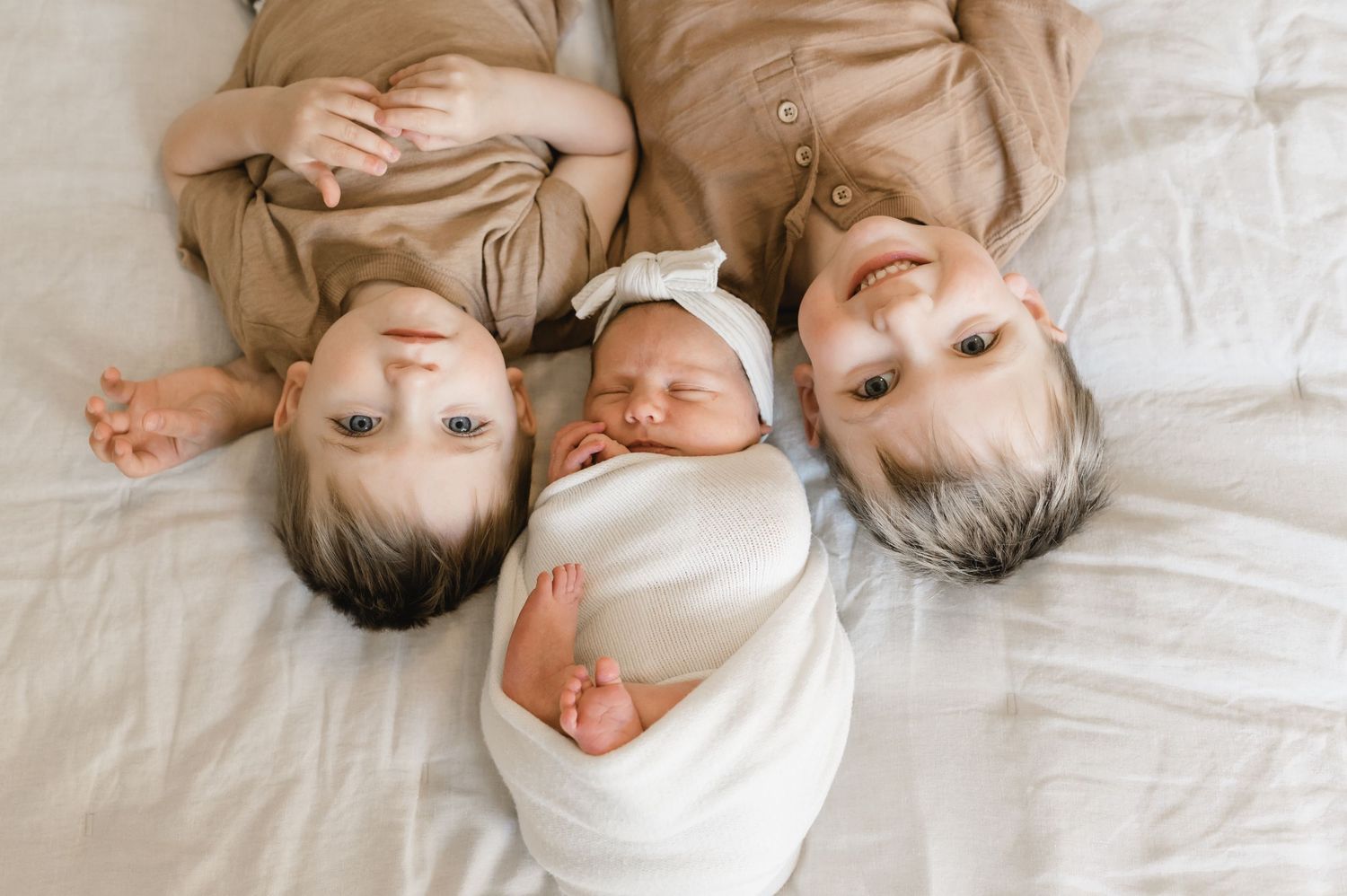 Two smiling siblings lying beside their swaddled newborn baby sister on a white bed. Wisconsin. Minnesota.