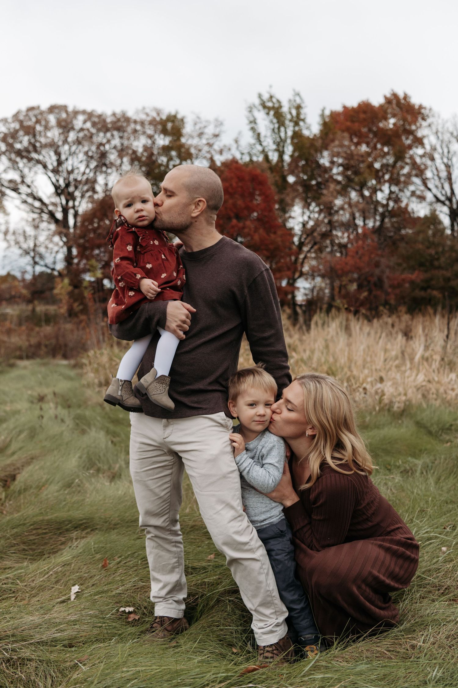 A family of four poses in an autumn field, parents kissing their two young children tenderly. Lake Elmo, Minnesota.