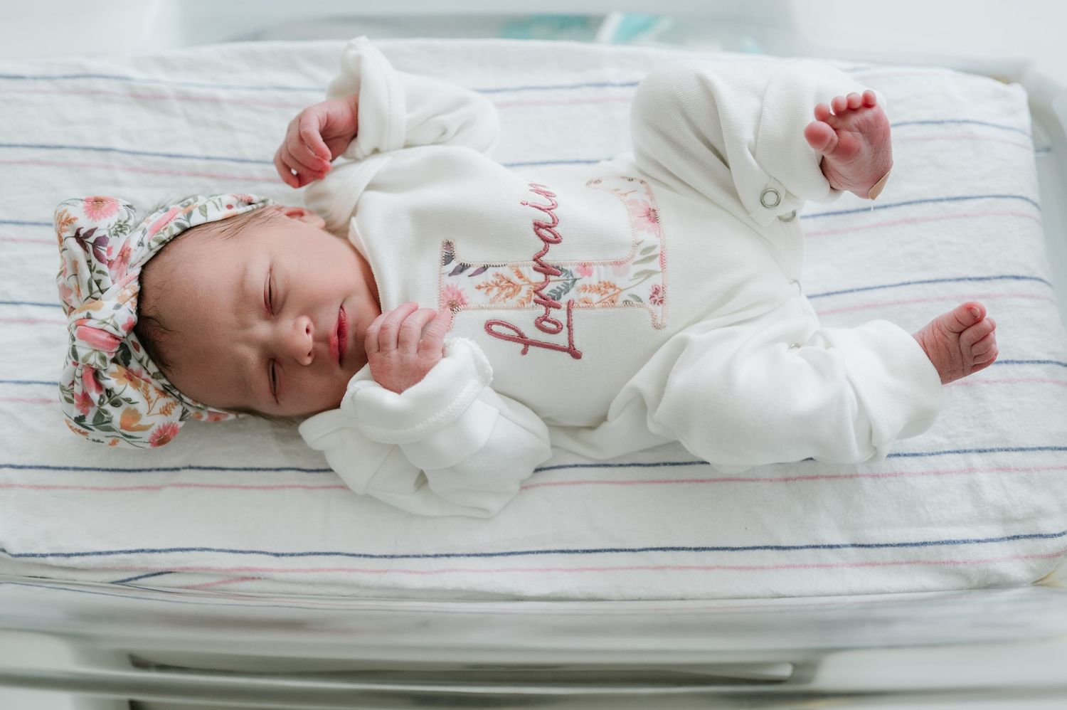 Sleeping newborn baby in personalized white outfit and floral headband in hospital bassinet. Fresh 48 hospital photos.