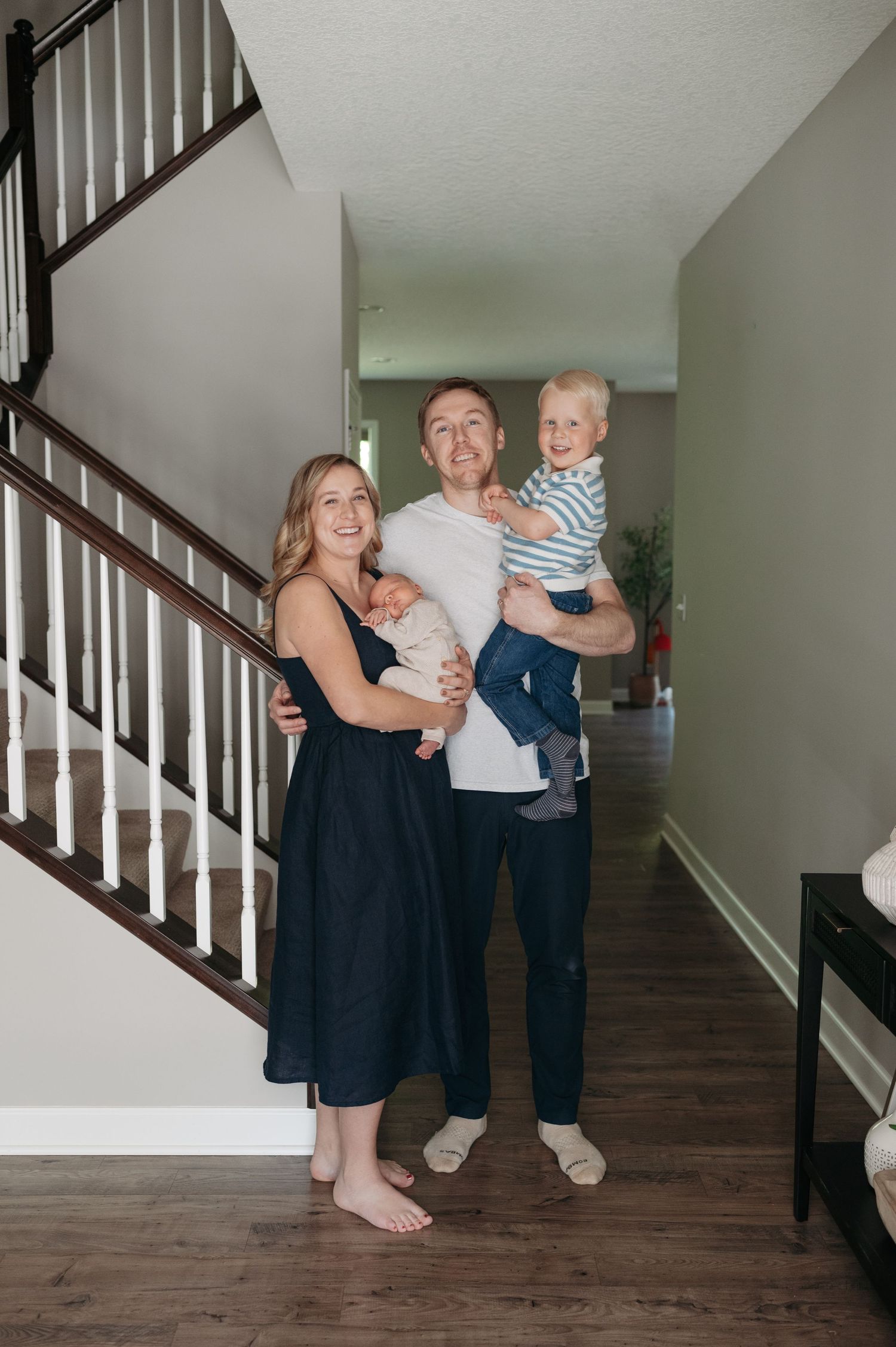 Happy family of four posing in home entryway with newborn and toddler by staircase.
