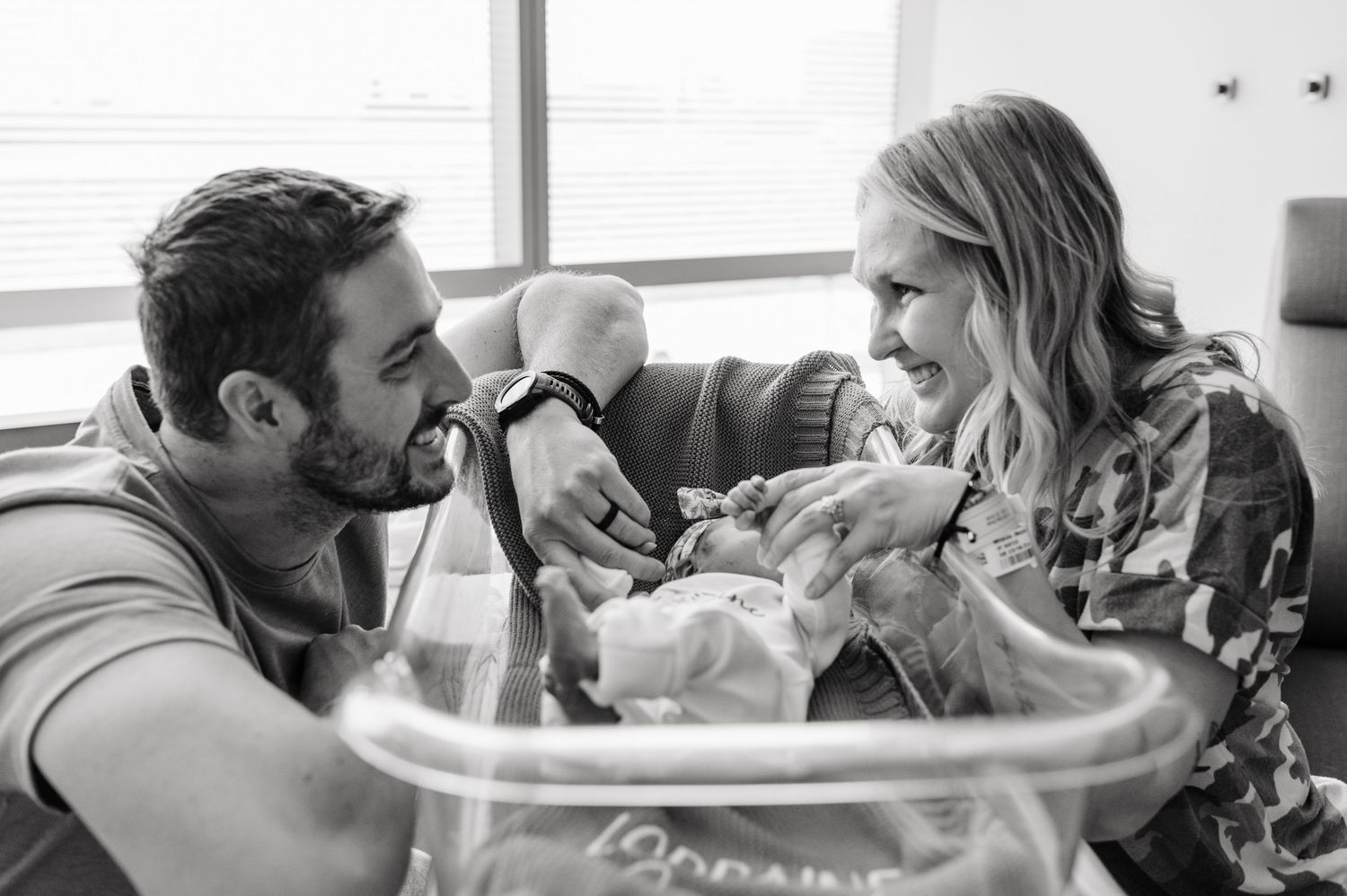 Black and white photo of new parents smiling at their newborn baby in hospital nursery. Fresh 48 hospital photos.