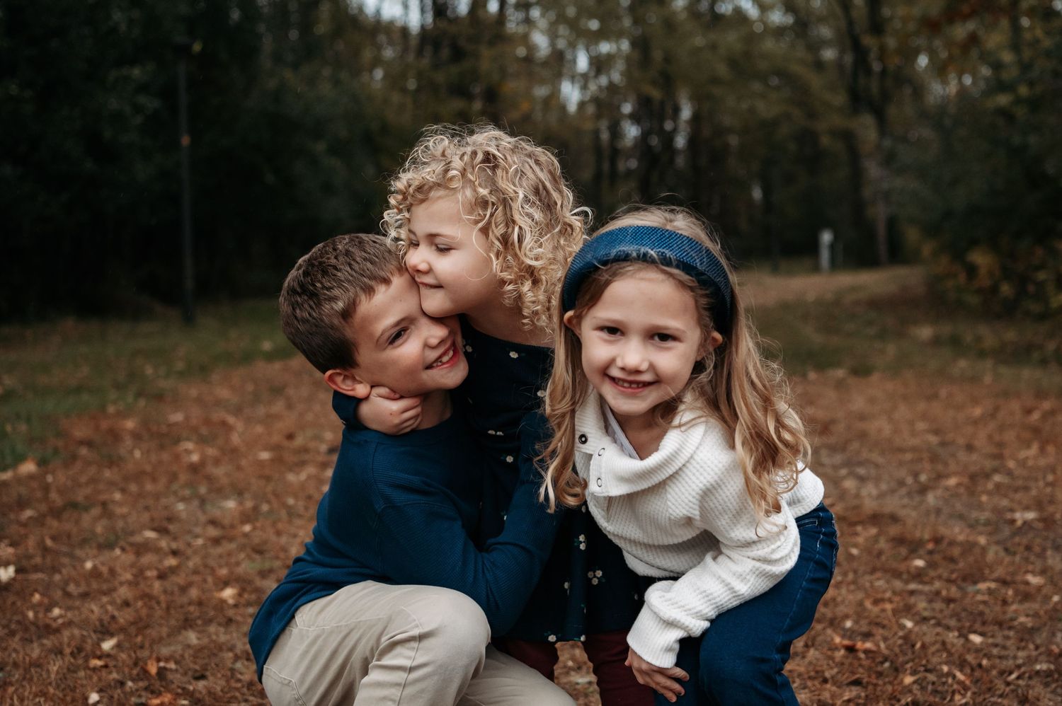 A father crouches in autumn leaves as two young daughters hug him from behind outdoors. Bloomington, Minnesota