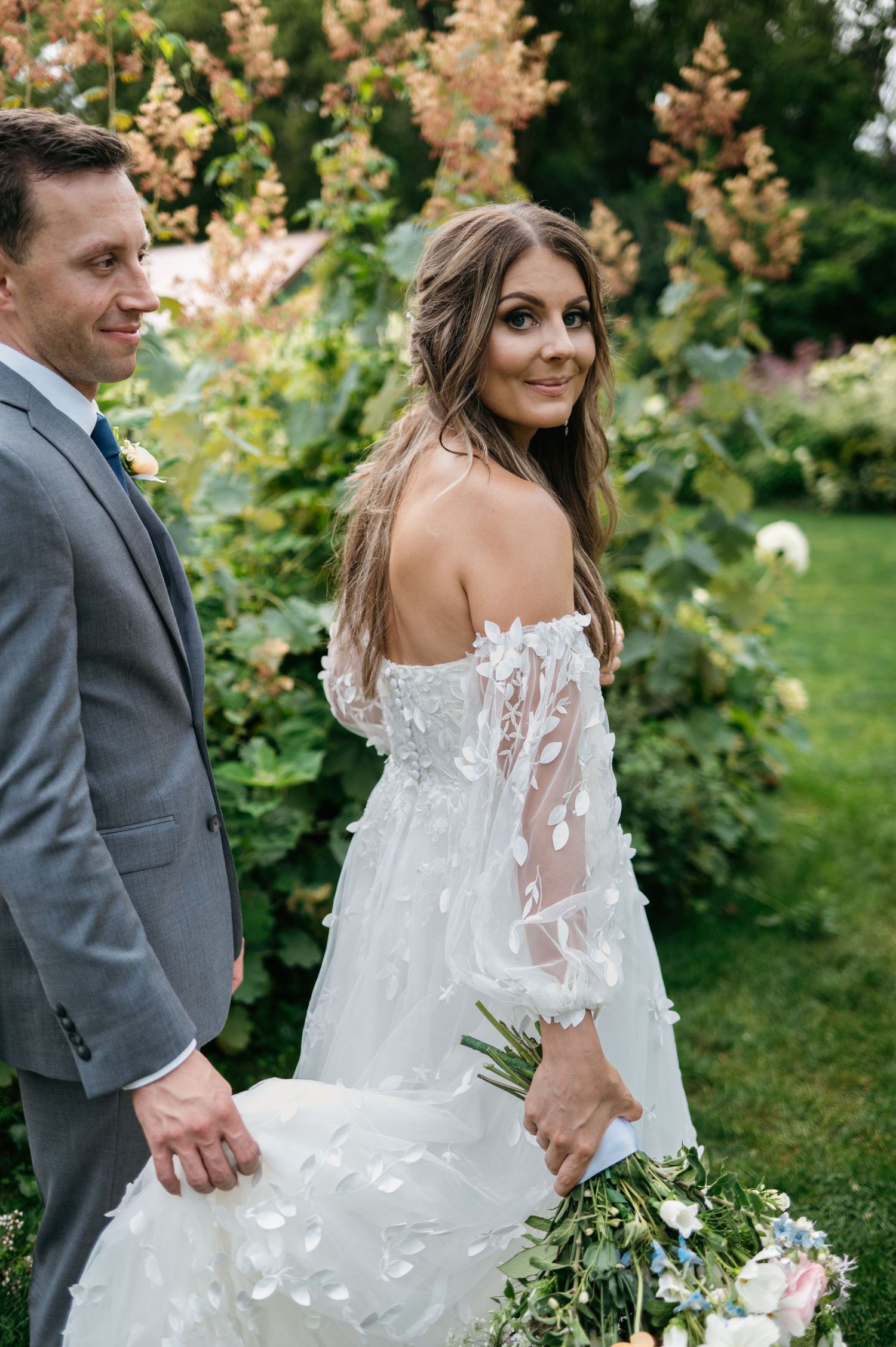 Bride in off-shoulder floral wedding gown smiling over shoulder holding bouquet in garden.