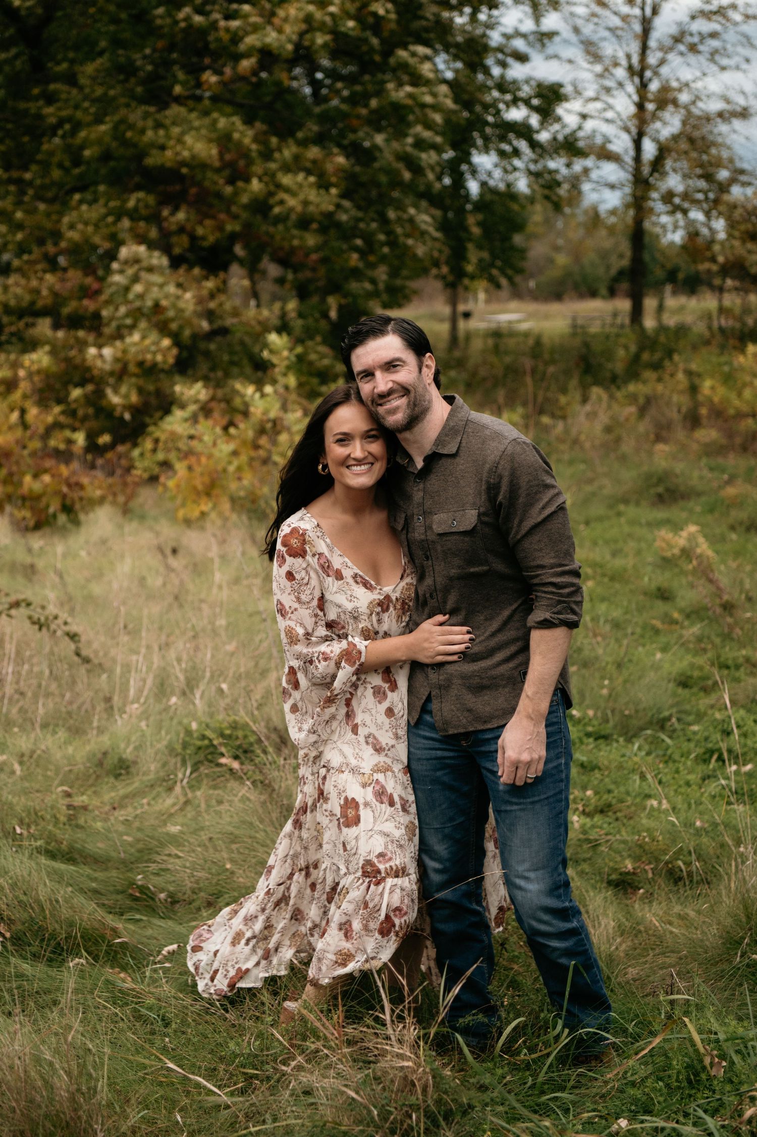 A smiling couple embraces in an autumn meadow, the woman wearing a floral dress and the man in a brown shirt. Woodbury, Minnesota.