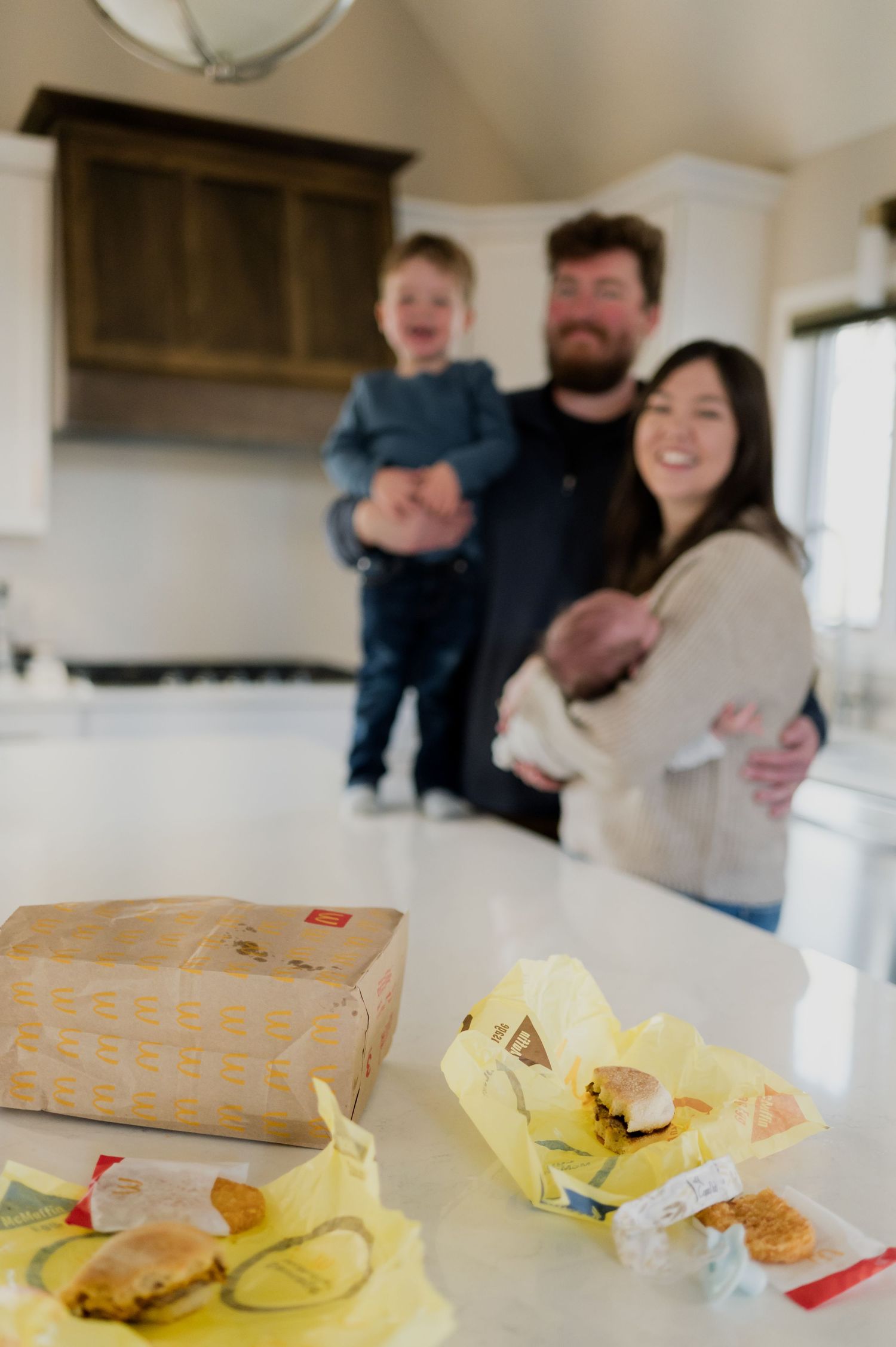 Happy family with toddler and newborn smiling behind McDonald's breakfast on a kitchen counter.