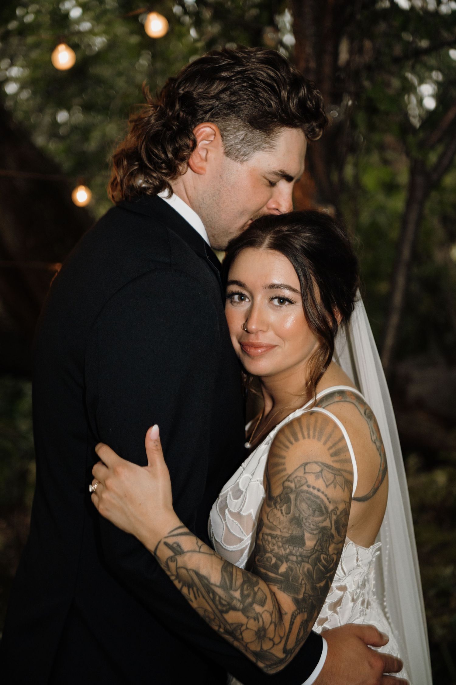 Bride with tattoo sleeve in lace gown embraces groom in black suit at outdoor evening wedding reception. Wisconsin. Minnesota.