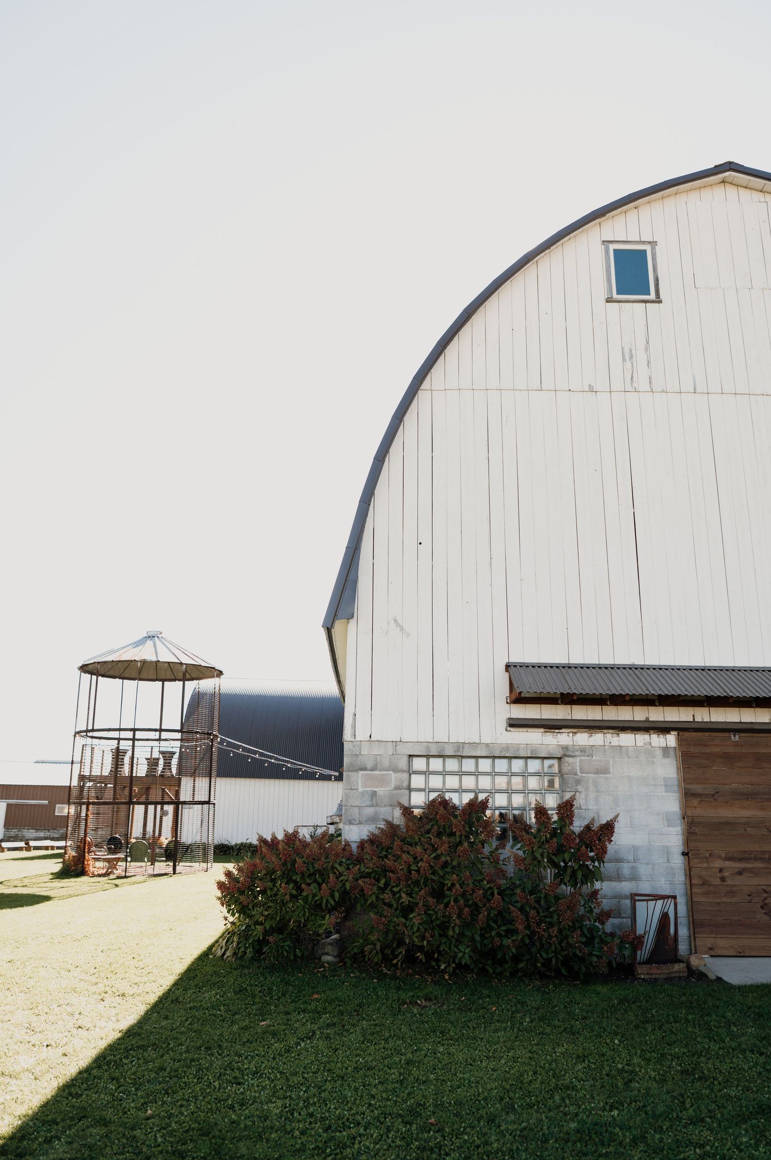 White barn wedding venue exterior with arched roof, stone foundation, and rustic wooden door surrounded by lush greenery.
