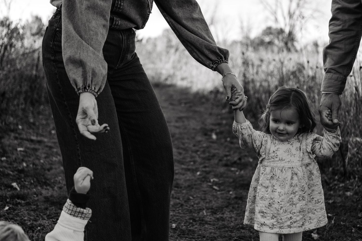 Black and white photo of toddler girl holding hands with two adults on a nature path. Woodbury, Minnesota.
