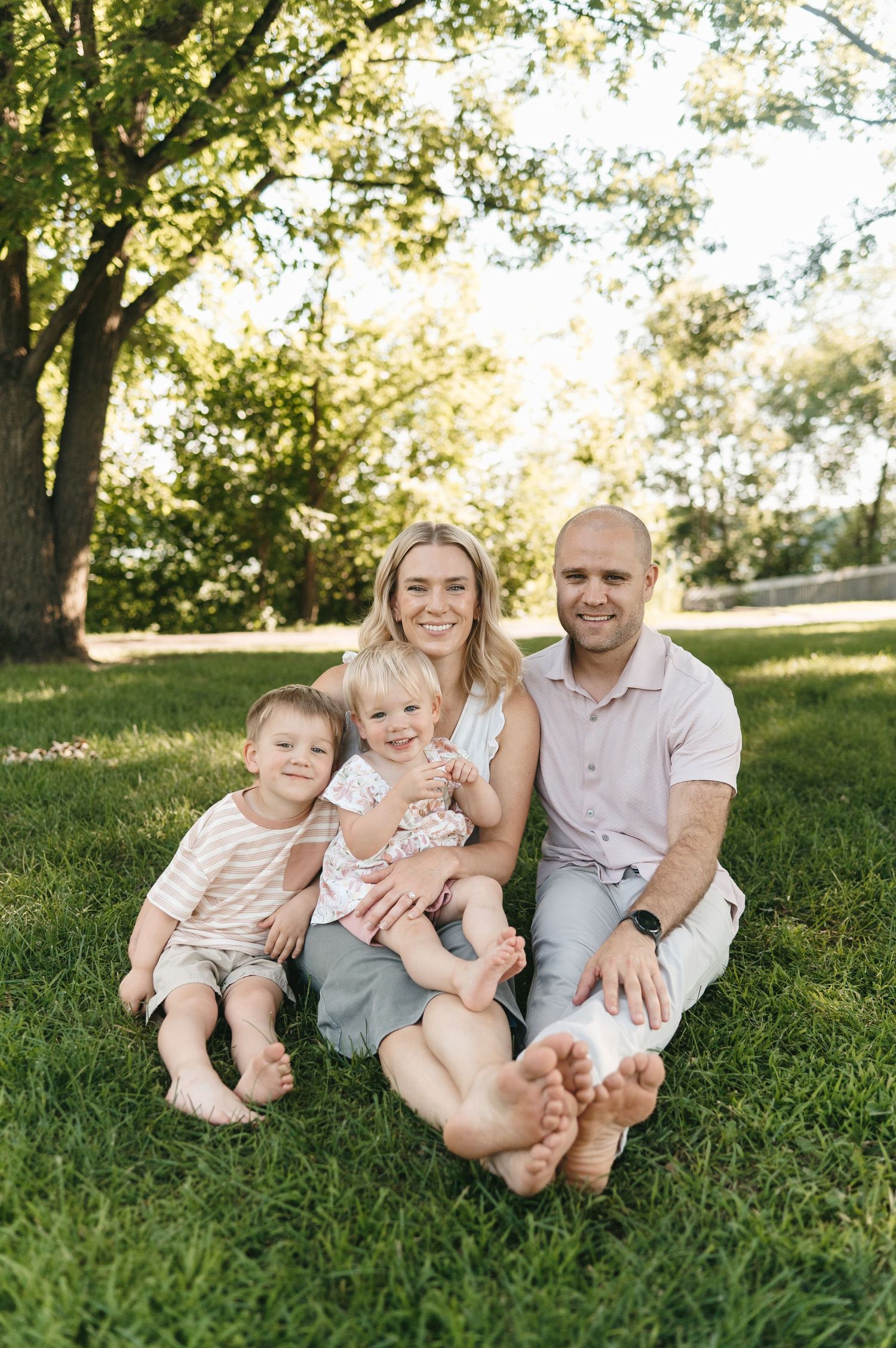 Happy family of four sitting barefoot on green grass under large tree in sunny park.
