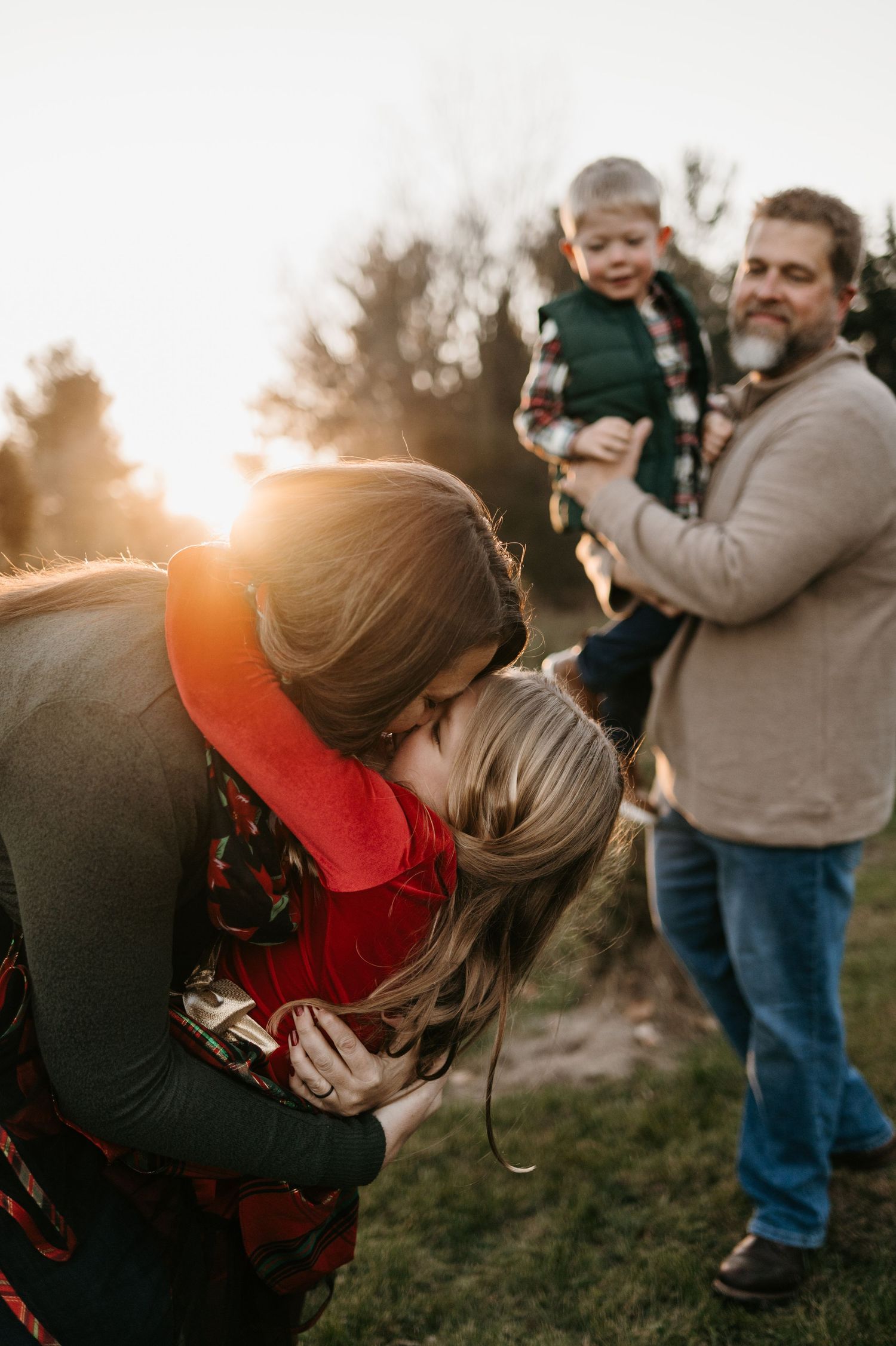 Mother kissing young daughter at golden sunset while father holds toddler son in background. Hudson Wisconsin