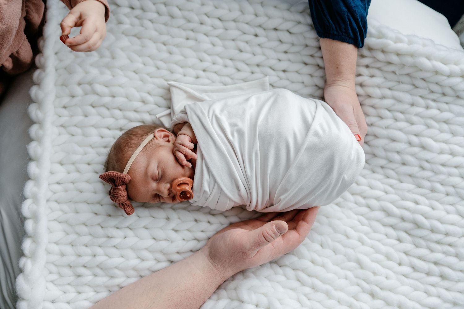 Swaddled newborn girl with pink bow headband lying peacefully on a white knit blanket.