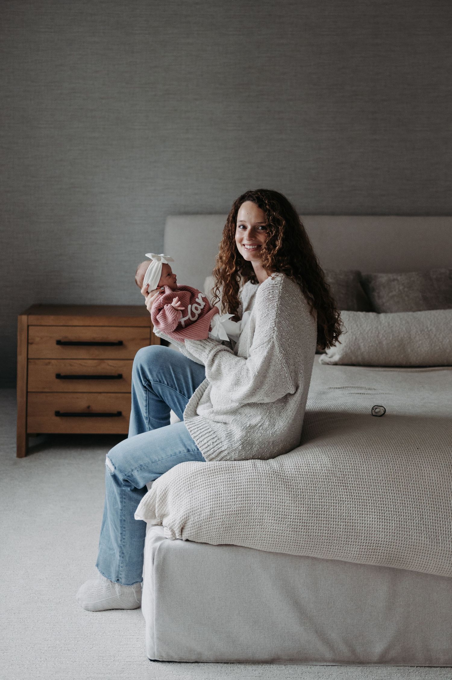 A smiling mother sits on a bed holding her newborn baby in a cozy bedroom.