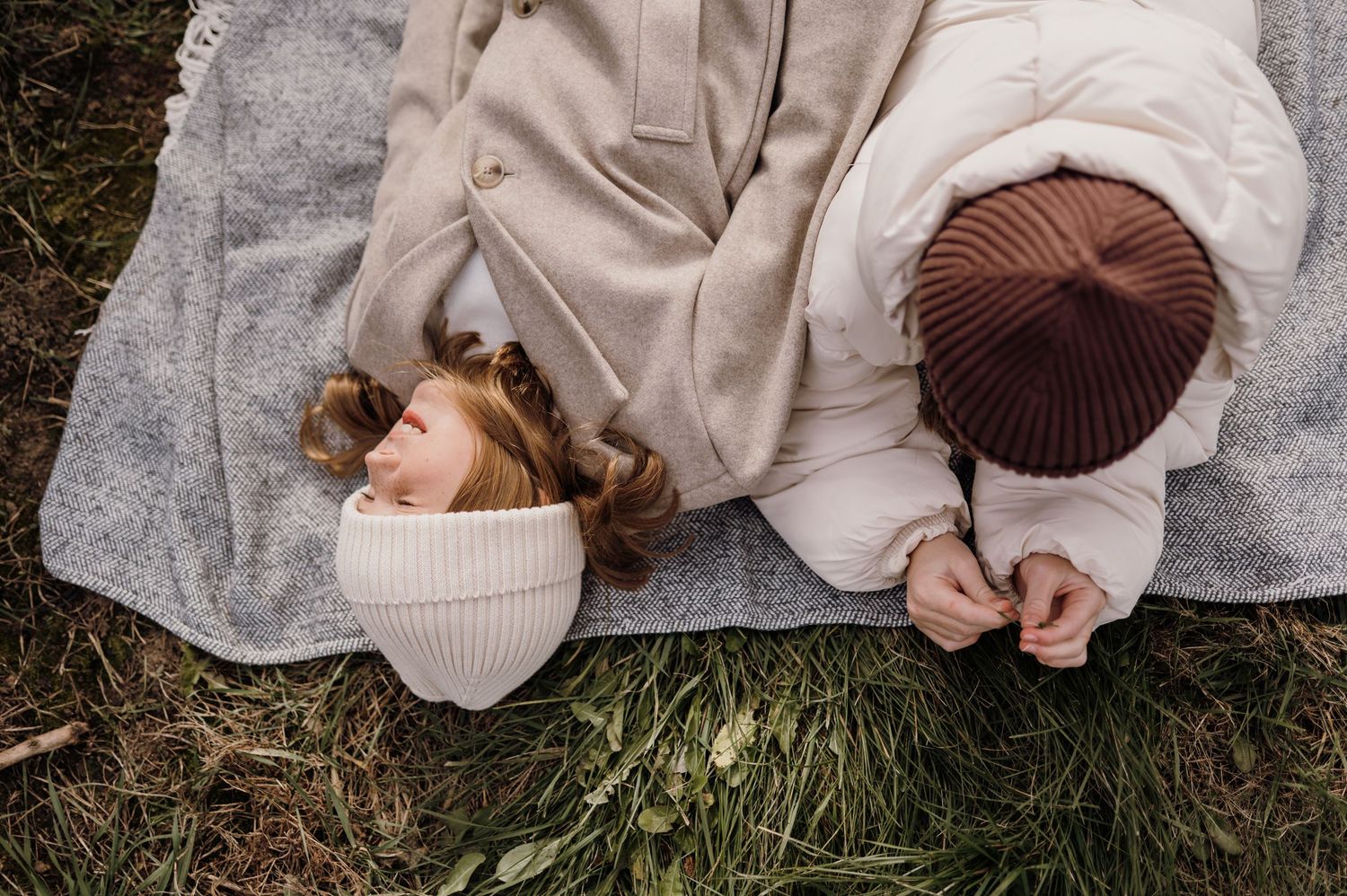 Two children lying on a blanket in grass, wearing winter hats, one holding a wildflower. Lake Elmo, Minnesota.