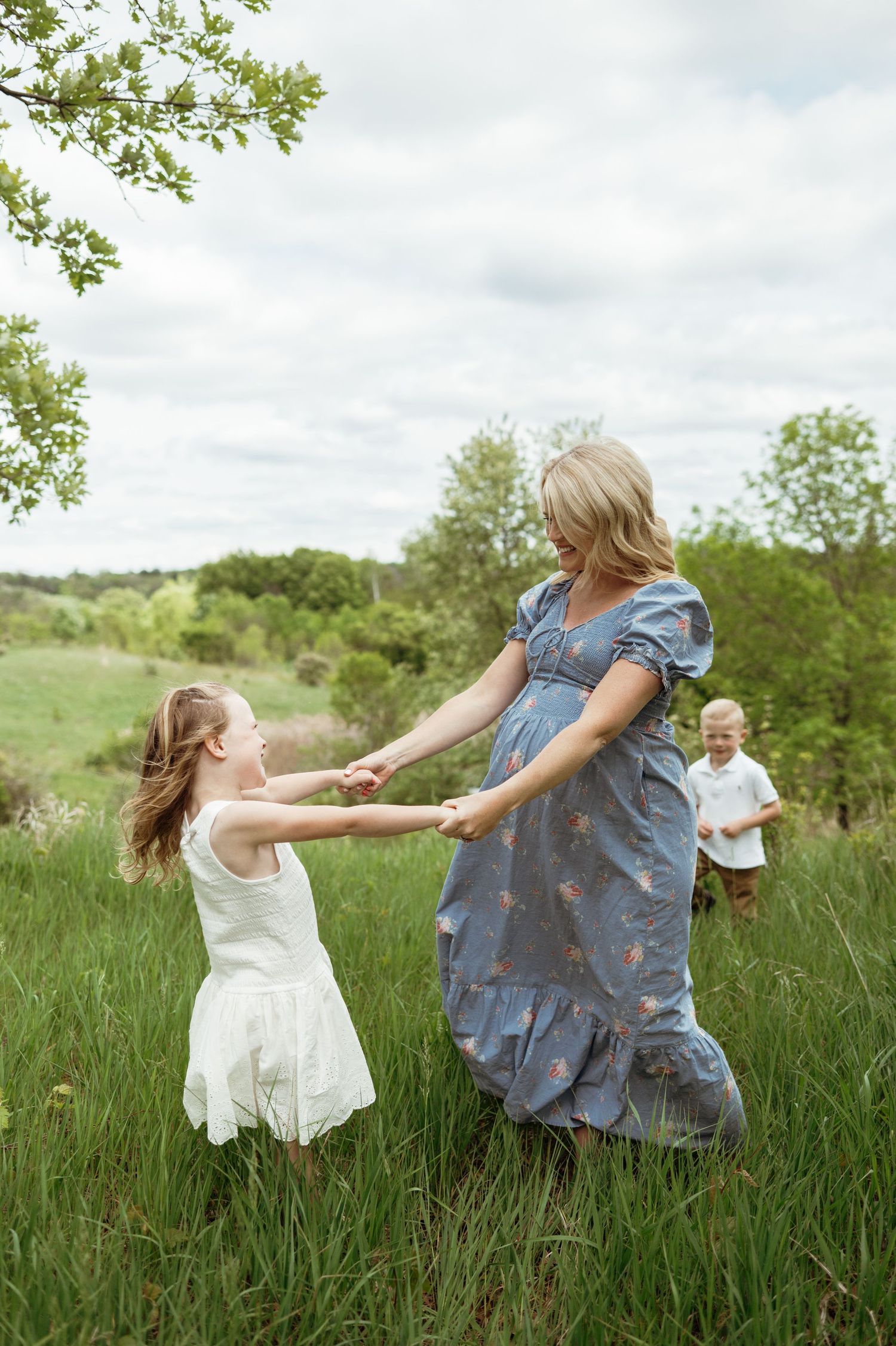 Pregnant mother in blue floral dress dancing with daughter in lush green summer field.