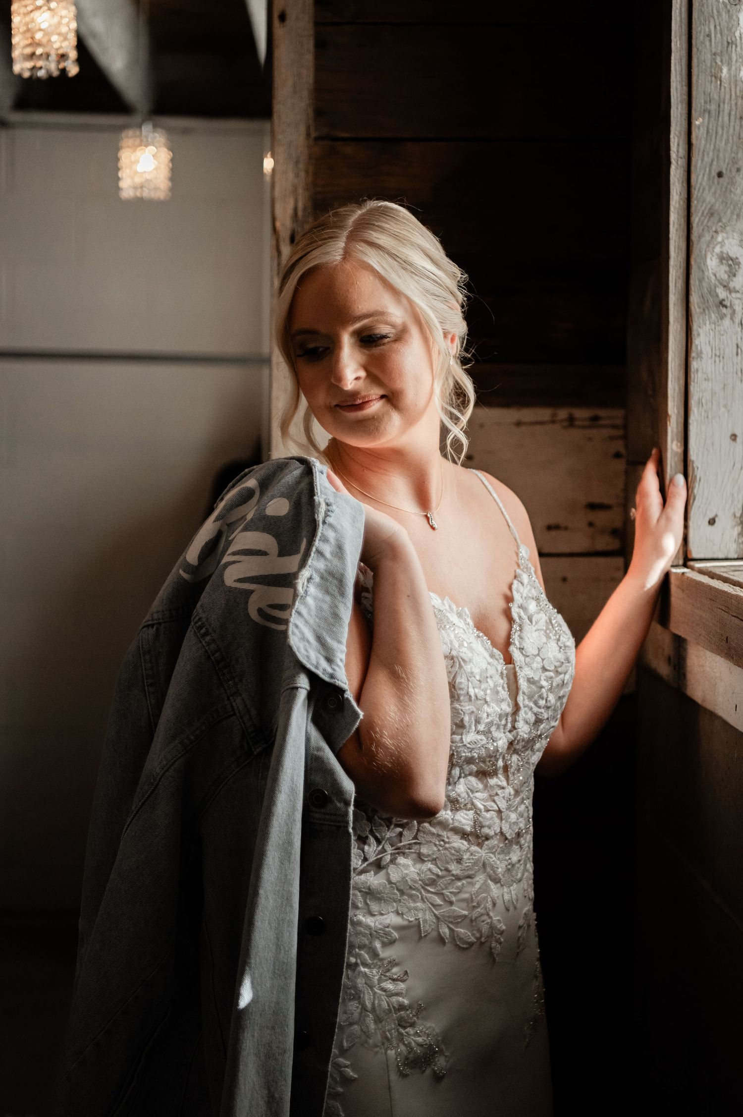 Elegant bride in lace gown holds jacket while posing in rustic barn doorway with warm lighting.