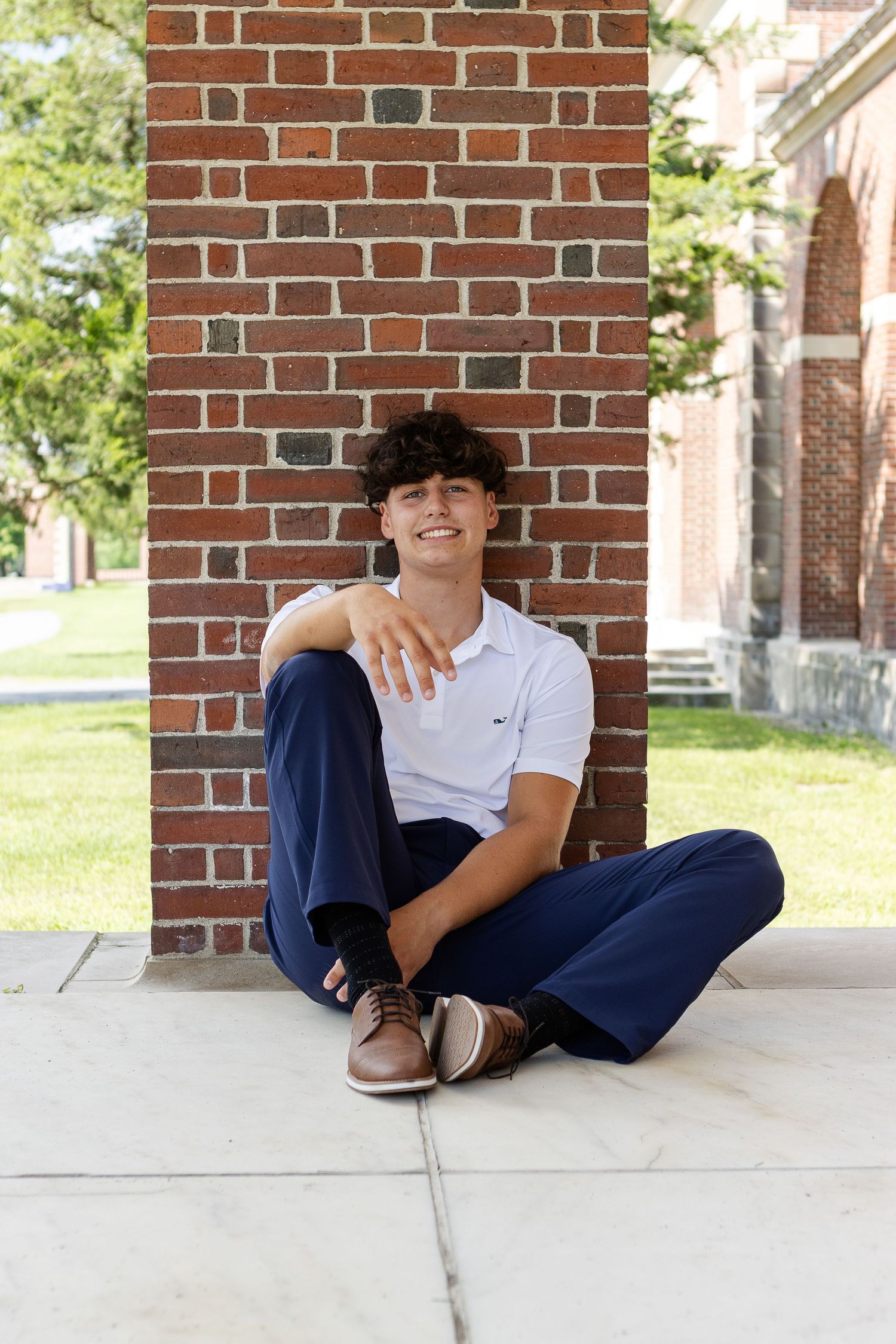 Smiling young man with curly hair sits on a concrete patio against a red brick pillar in a white polo, navy pants and brown shoes.